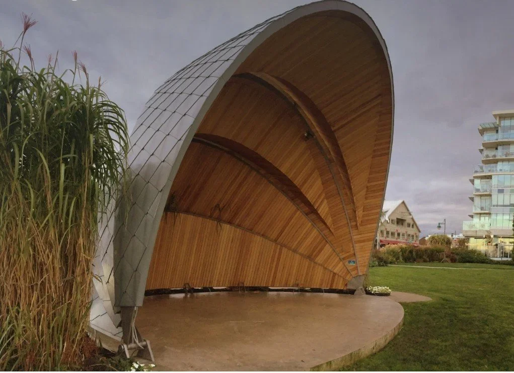 Modern outdoor stage with wooden interior and curved metal roof over green lawn, neighboring building in background.