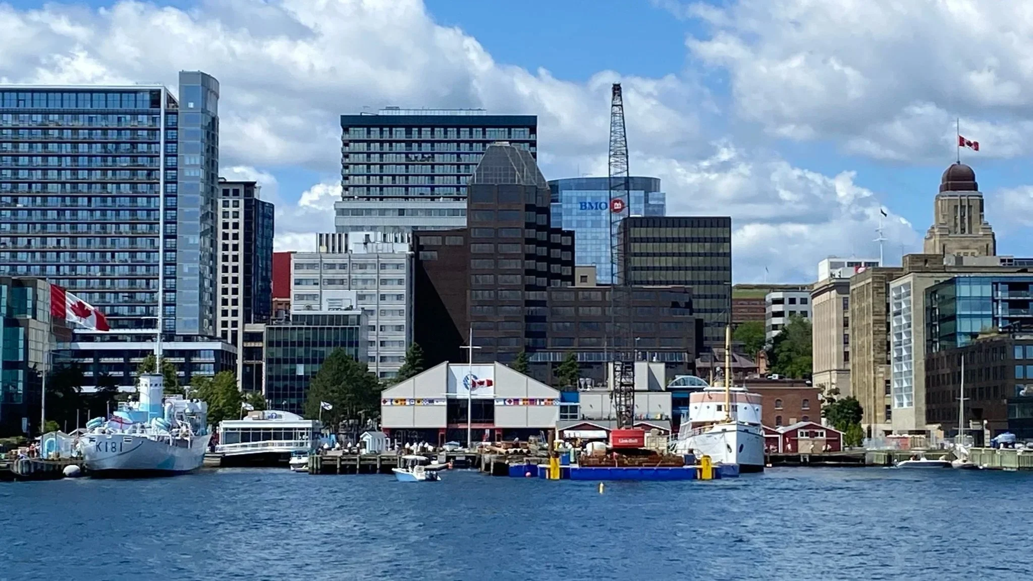 City skyline with tall buildings, boats docked at a waterfront, a Canadian flag, and partly cloudy blue sky.