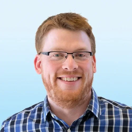 A smiling man with red hair, glasses, and a beard, wearing a blue plaid shirt, against a light blue background.