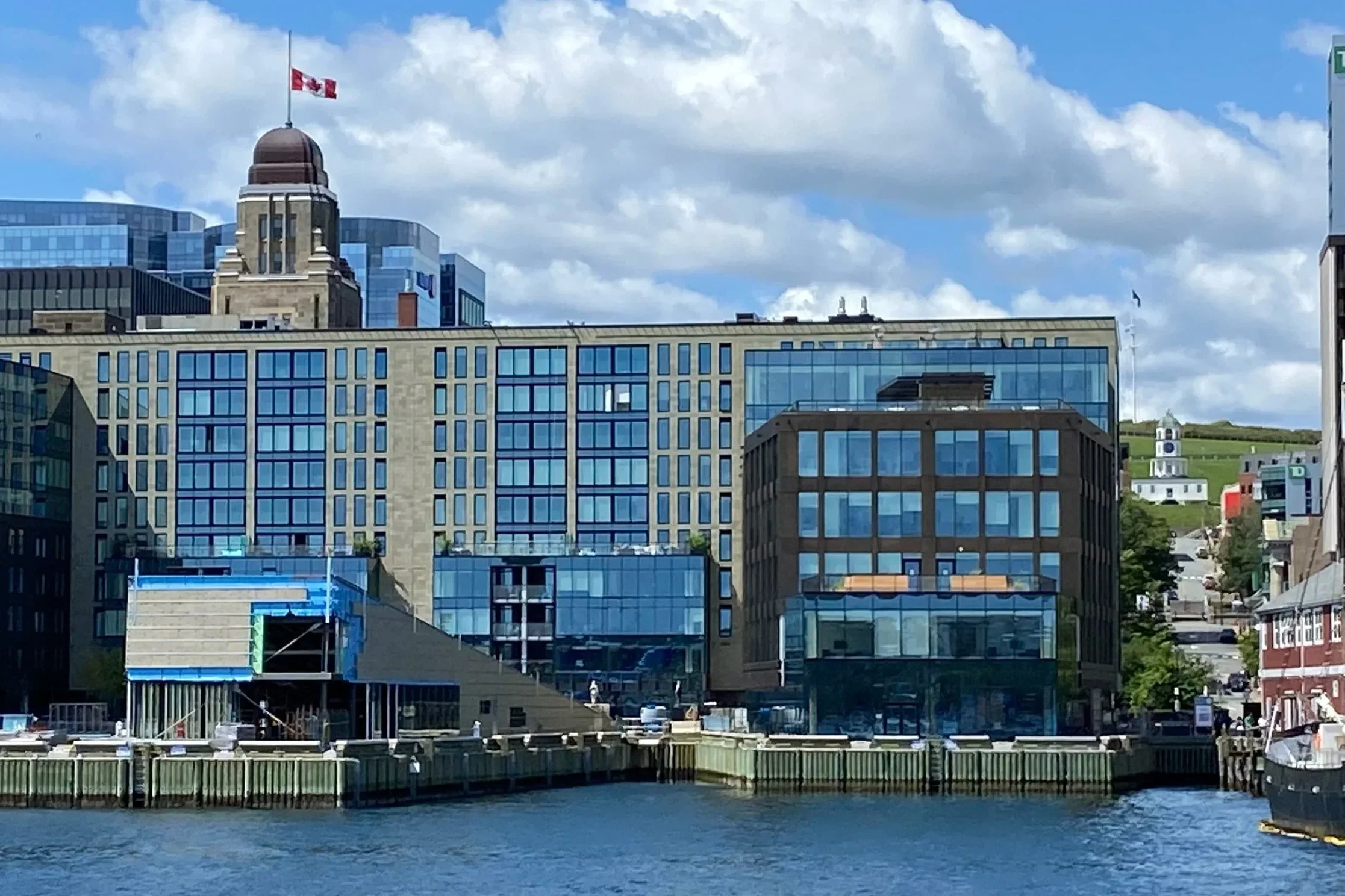 Cityscape with modern glass buildings along water, a hill with a historic church and flag in the background, and partly cloudy sky.