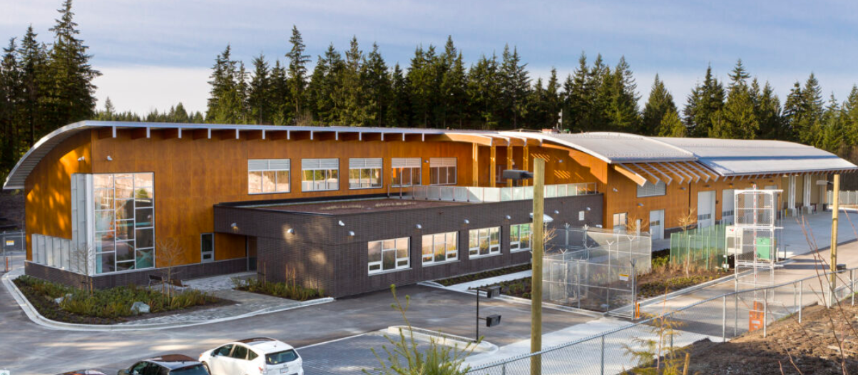 Modern school building with curved roof and large windows, surrounded by fencing and parking lot, set against a background of trees and a cloudy sky.