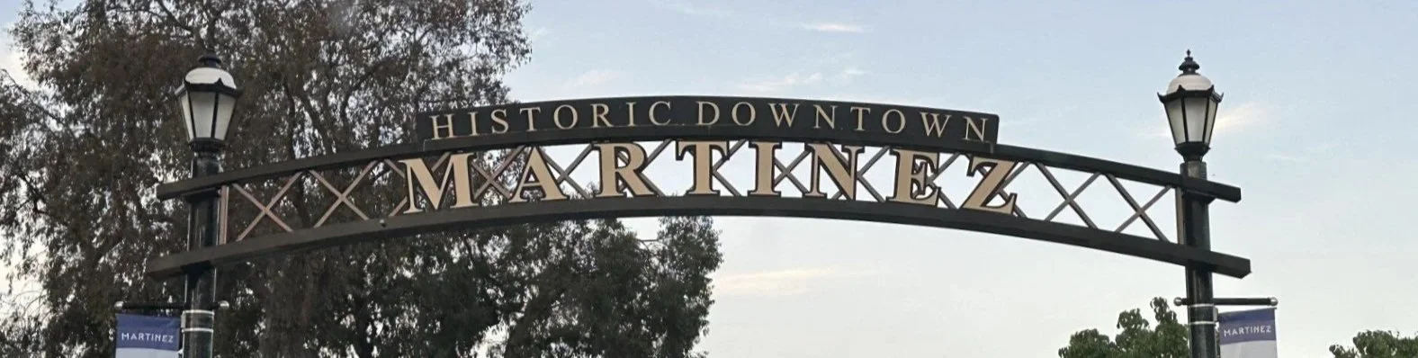 An archway sign reading "HISTORIC DOWNTOWN MARTINEZ" in a city park or street, with trees and sky in the background.