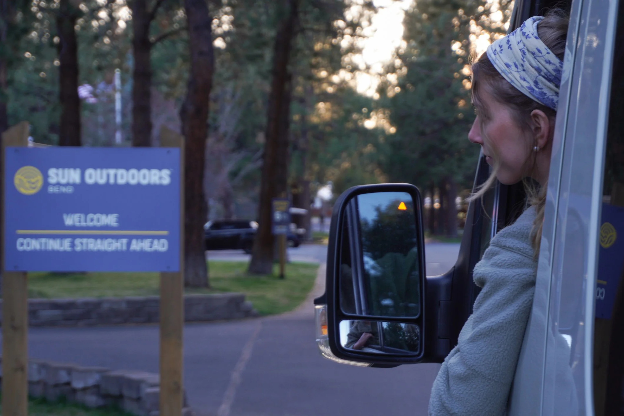 A woman in a light-colored jacket and a headscarf looking out from the side door of a vehicle at a park entrance sign that reads 'Sun Outdoors Bend. Welcome, Continue Straight Ahead' during sunset.