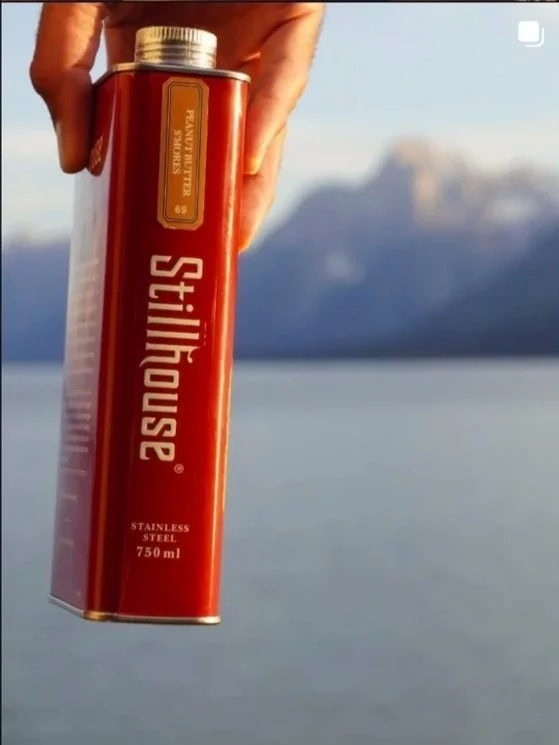 A hand holding a rectangular red stainless steel canister with a silver cap, featuring the brand name 'Stillhouse' and measuring 750 ml, against a blurred outdoor landscape background.