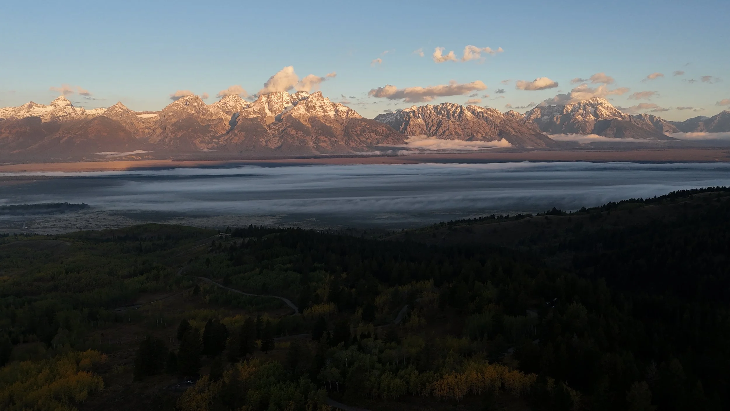 Snow-capped Grand Teton in the background with clouds around the peaks, a body of water in the middle, and forested hills in the foreground at sunrise.