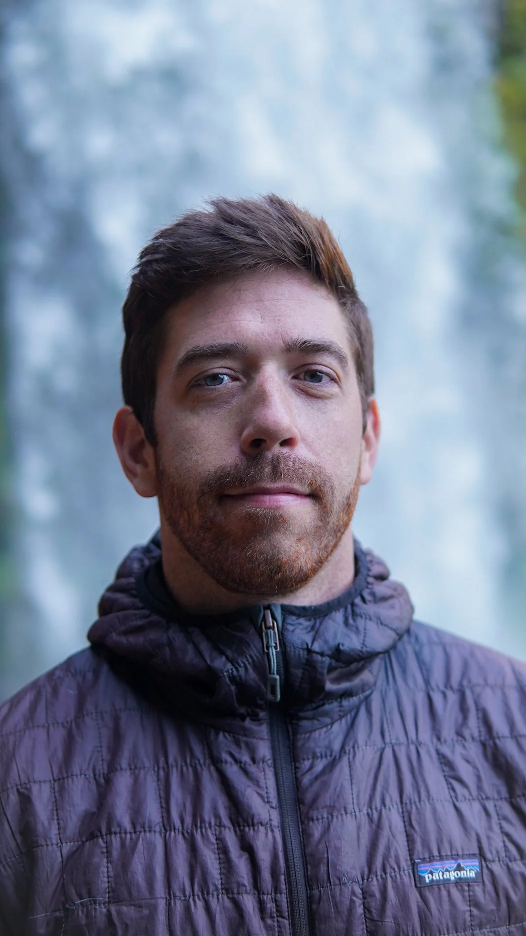 A man with brown hair and a beard wearing a dark purple Patagonia jacket standing outdoors with a misty forest background.