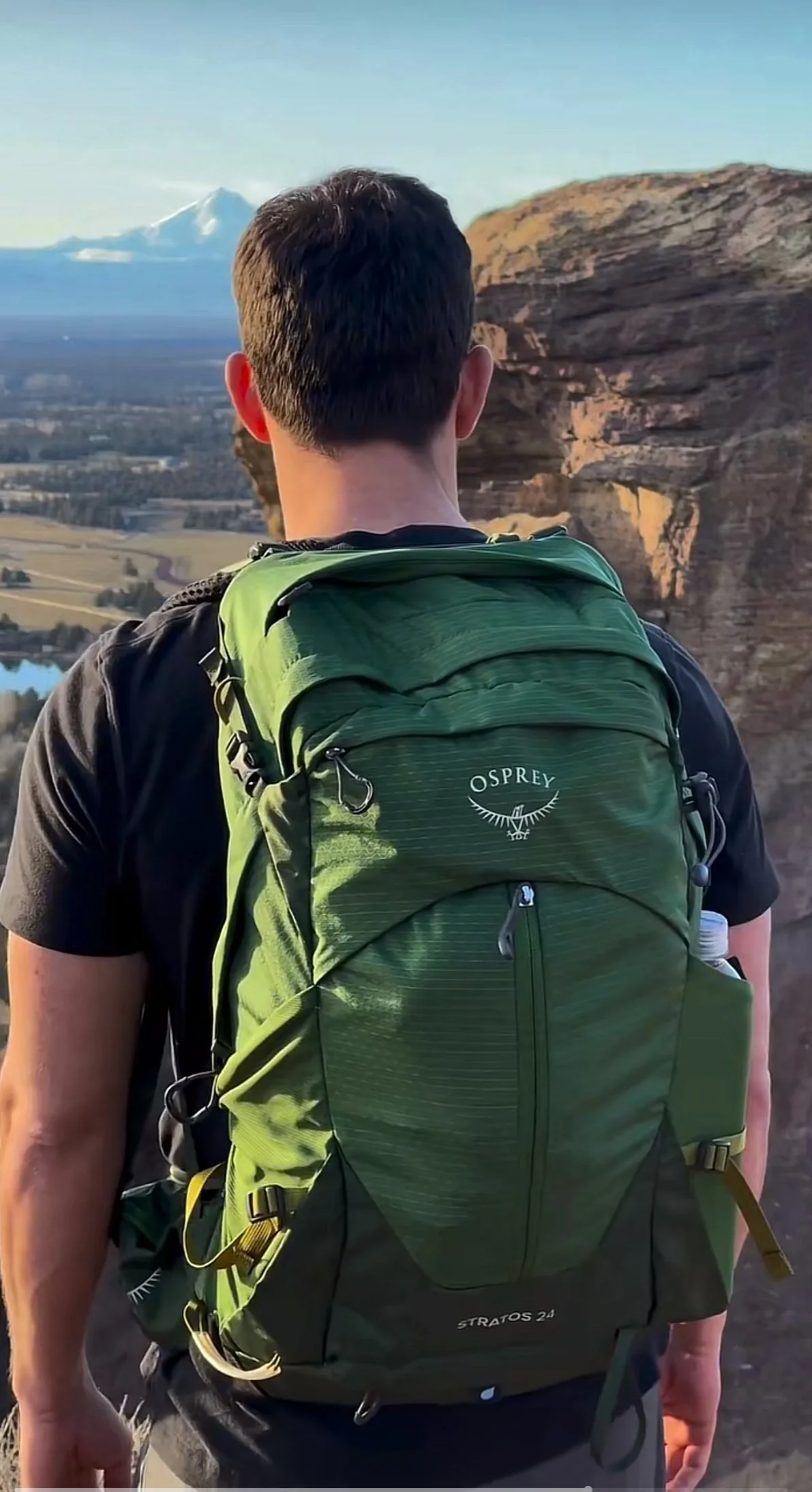 Man with a green Osprey backpack standing outdoors at Smith Rock State Park, looking at a landscape with a mountain in the background.