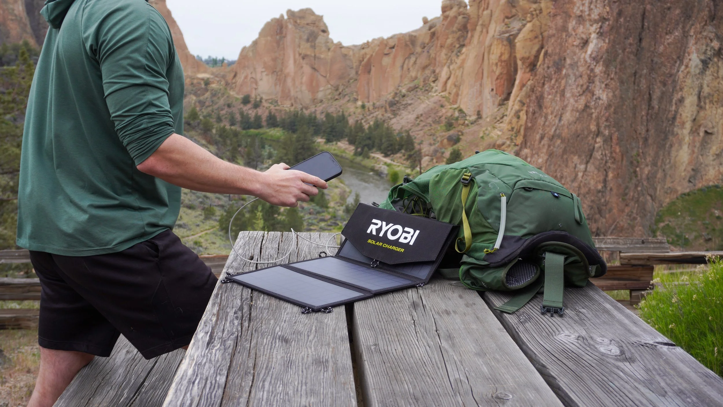 A person in a green shirt and black shorts using a smartphone outdoors near a wooden picnic table with a green backpack, a Ryobi solar charger, and a portable solar panel, with a river and rock formations in the background.
