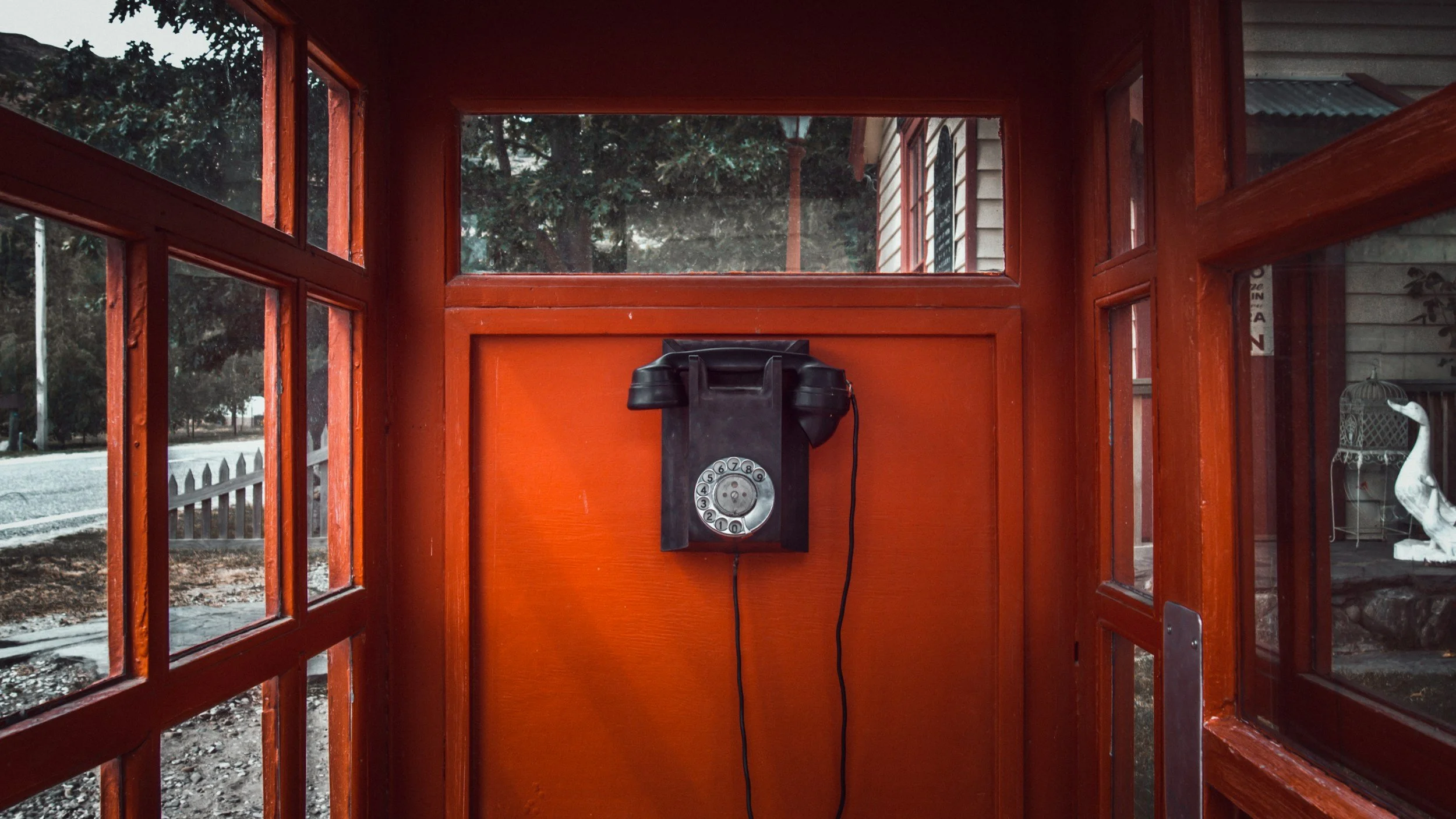 A vintage black rotary dial telephone mounted on the orange-painted interior wall of a small wooden booth with windows.