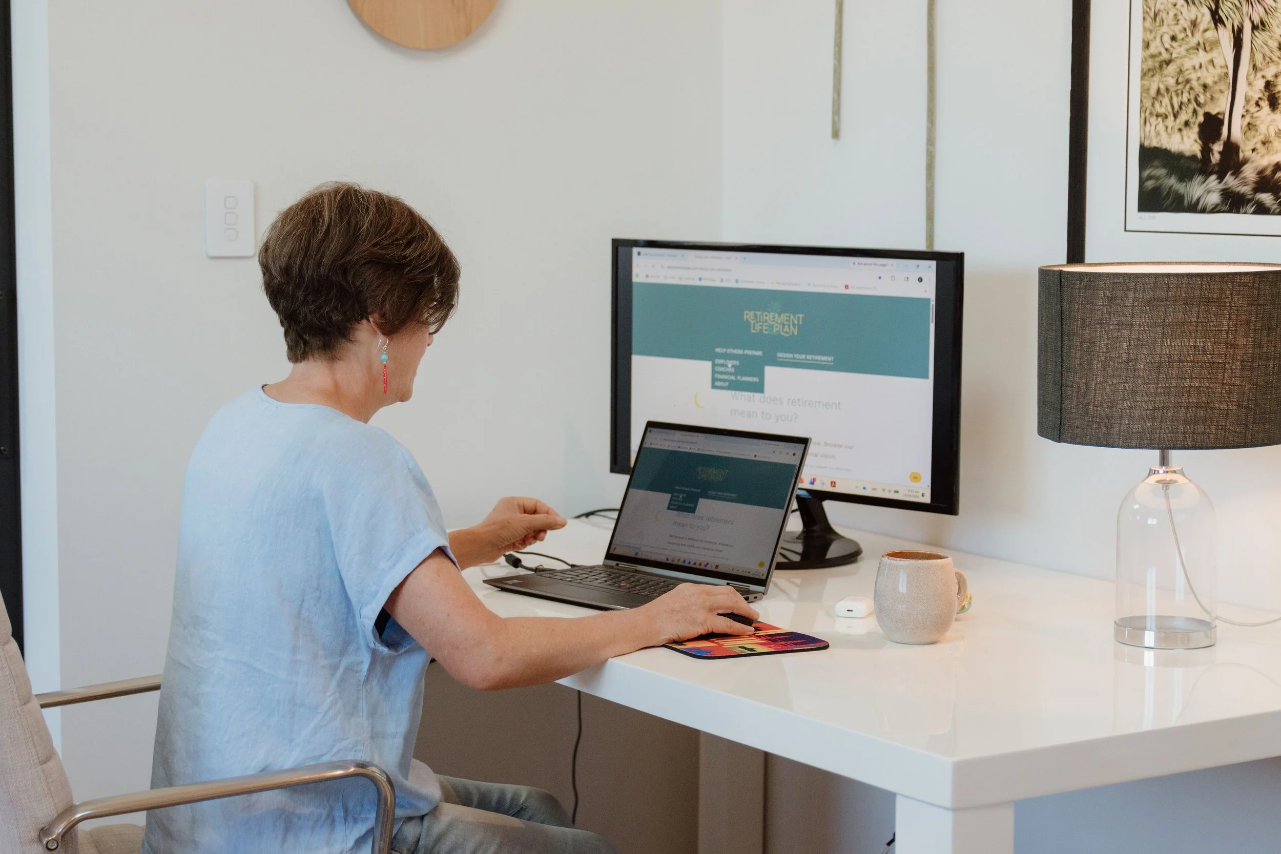 A woman with short brown hair, wearing a light blue shirt and colorful earrings, sitting at a white desk and looking at a laptop. On the desk, there is a large monitor displaying a retirement plan webpage, a small tablet, a beige reusable mug, a white wireless mouse, and a tall gray lamp. The background includes a black and white art print of a dog, white walls, and a round wooden wall decor.