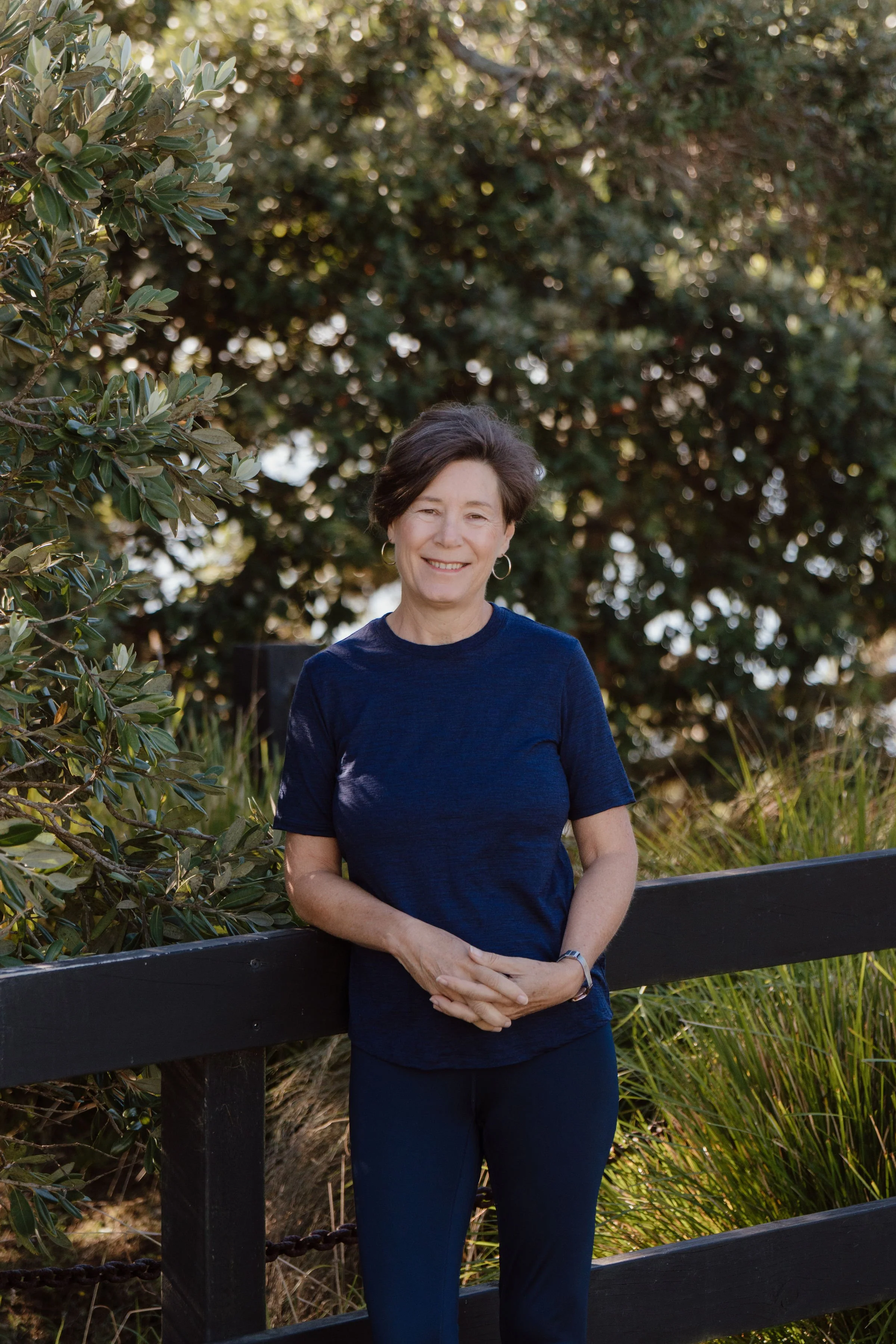 A woman with short dark hair, wearing a navy blue shirt and dark pants, standing outdoors in front of green foliage and a black wooden fence, smiling at the camera.