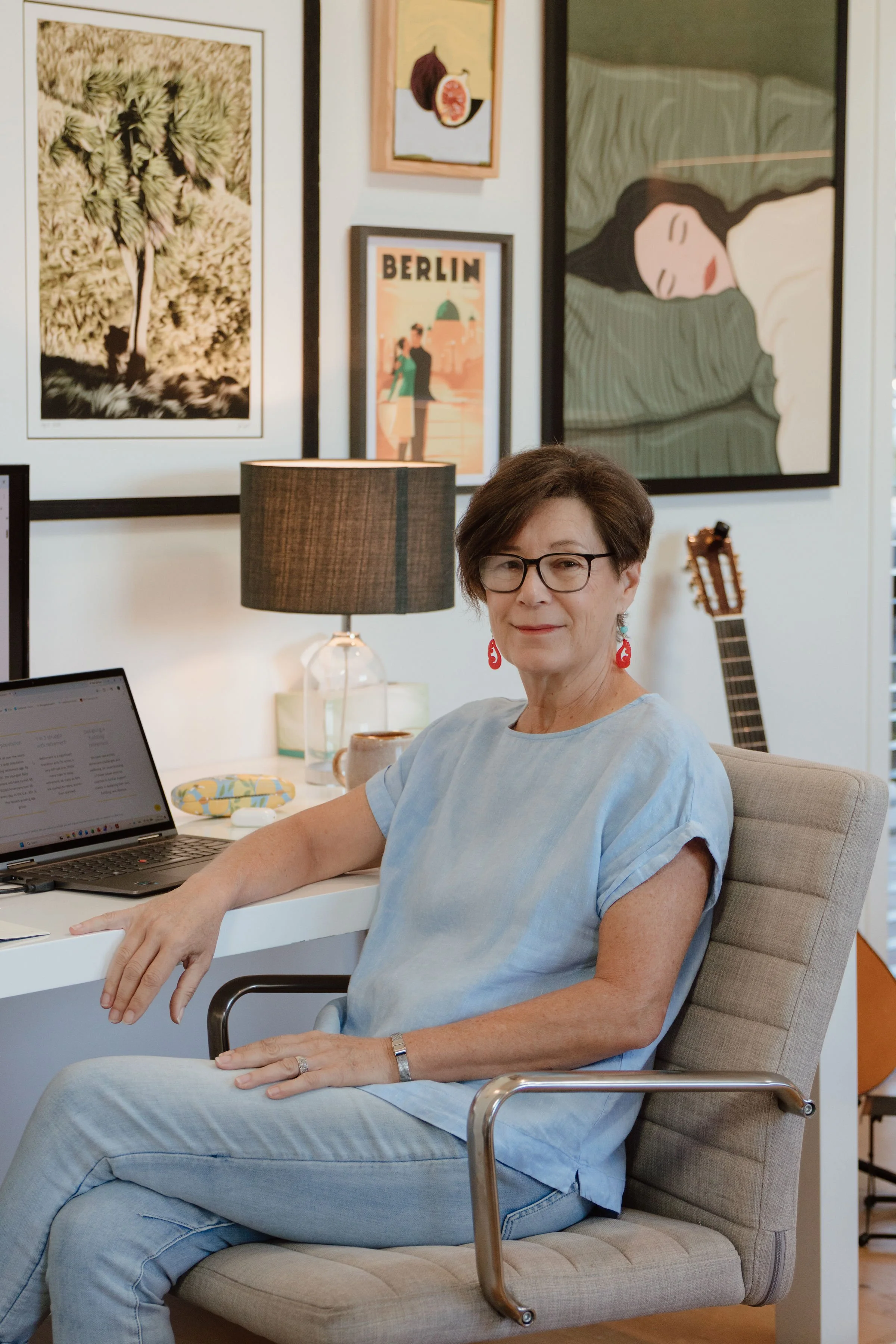 A middle-aged woman with short brown hair, glasses, and earrings sitting in a gray office chair at a white desk. She is wearing a light blue top and light pants. On the desk there is a laptop, a coffee mug, and a yellow and blue patterned item. She is in a room decorated with framed artwork on the wall, including a black and white tree drawing, a poster with the word "Berlin," and an abstract portrait of a sleeping woman. There is a lamp with a dark shade and a guitar in the background.