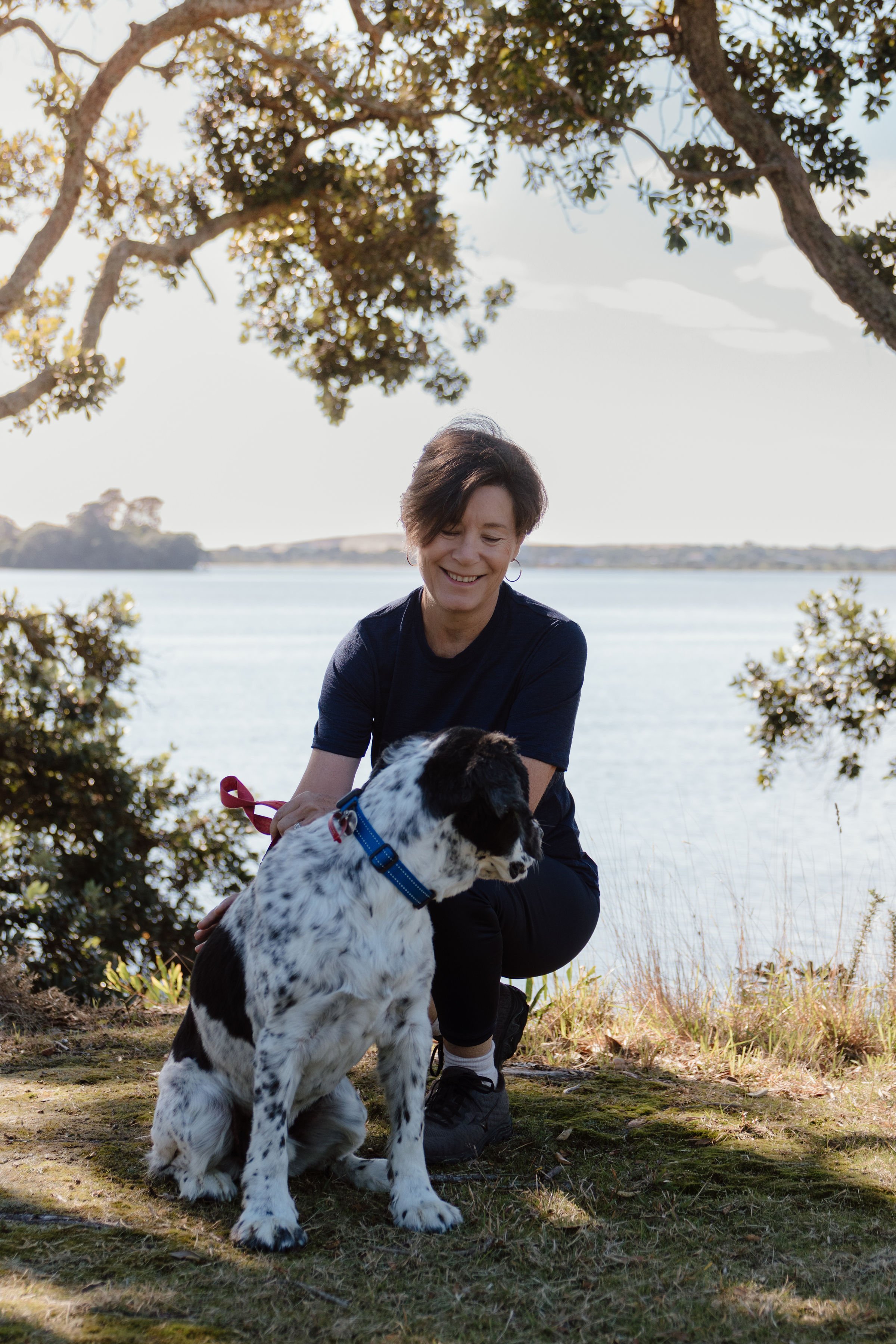 A woman kneeling outdoors on grass, smiling at a black and white spotted dog with a blue collar, near trees with a body of water in the background.