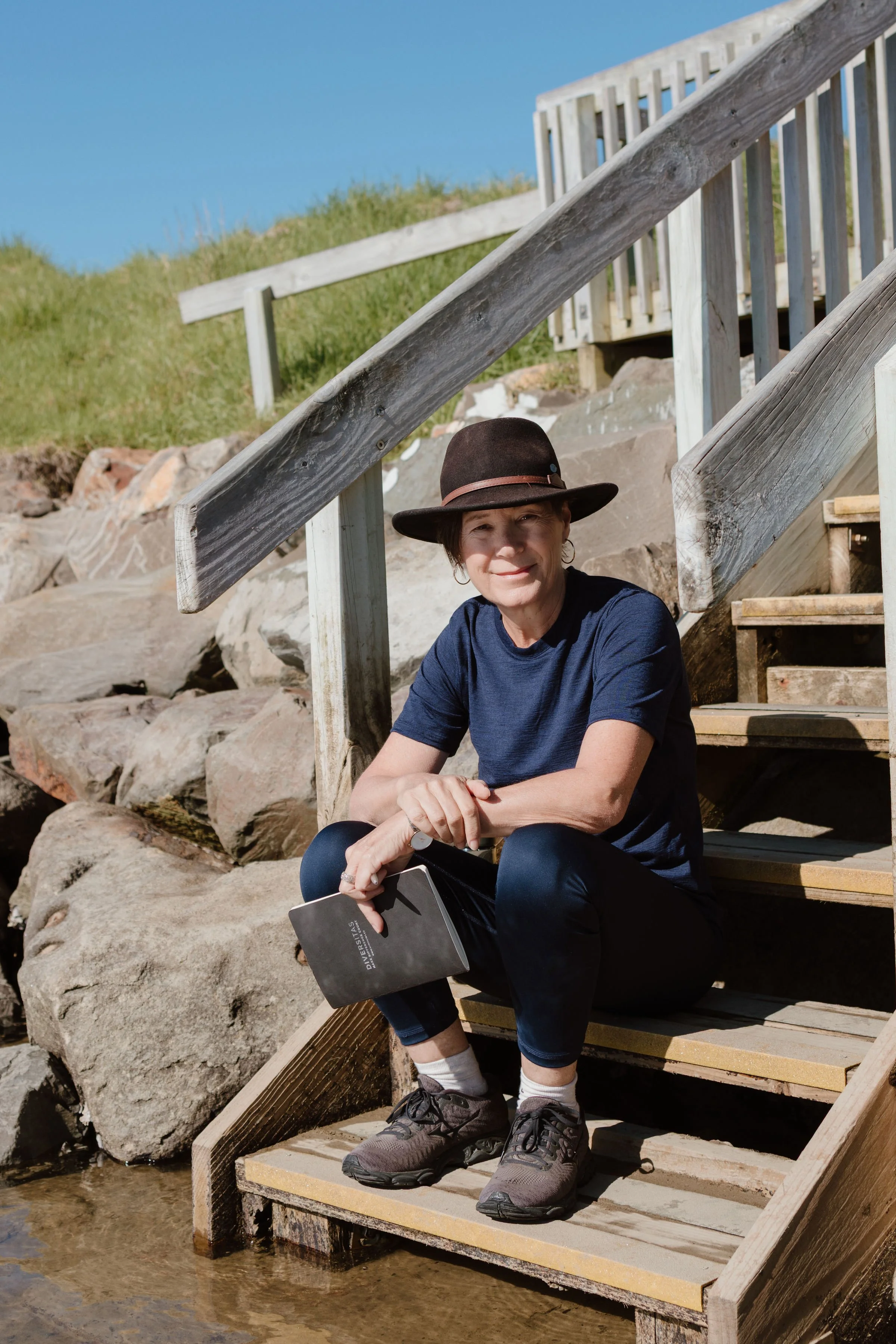 A woman wearing a brown wide-brimmed hat, navy blue shirt, black pants, and hiking shoes sitting on wooden stairs next to water. She is smiling and holding a notebook. Stone rocks and a grassy hill are in the background.