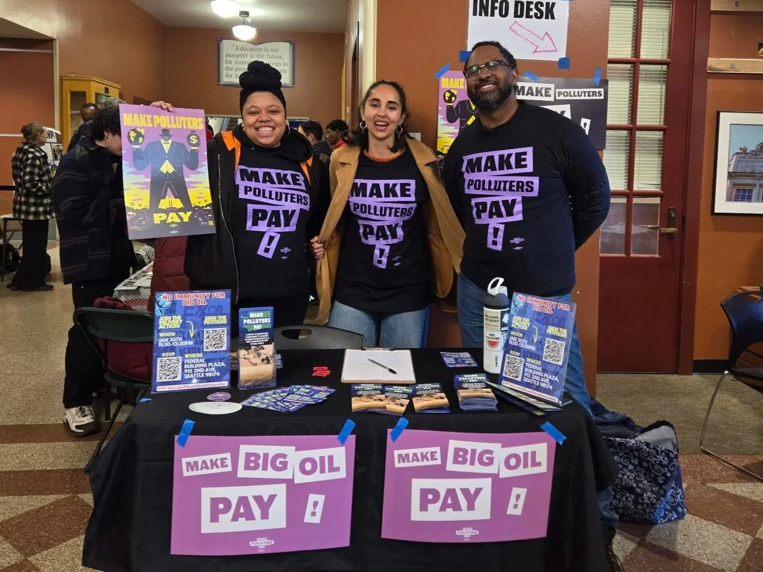 Three people standing behind a table at an event, wearing black T-shirts that say 'MAKE POLLUTERS PAY!', smiling for the photo. The table has signs that say 'MAKE BIG OIL PAY!' and informational flyers.