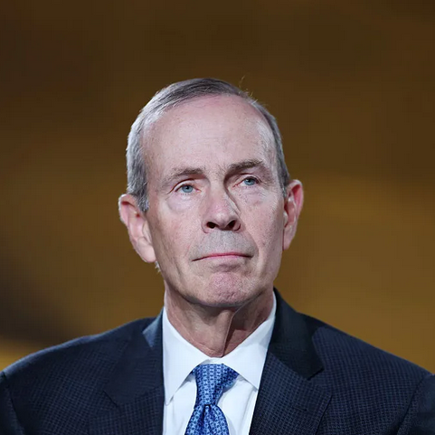 A middle-aged man in a suit and tie with a serious expression, against a plain dark background.