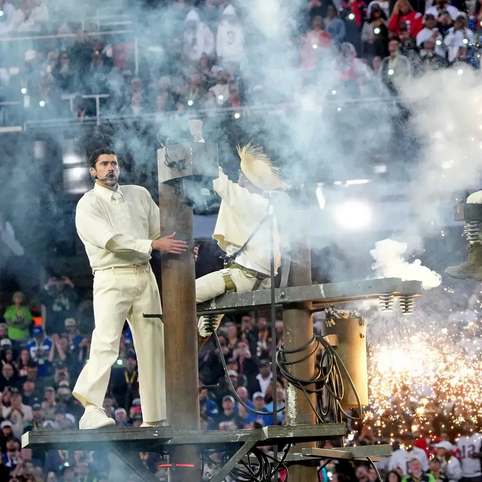 Person in white outfit standing on a platform amid fireworks and smoke, with a crowd in the background.