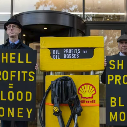 Protesters holding signs criticizing oil profits in front of a Shell gas station, with a yellow shell fuel pump in the background.