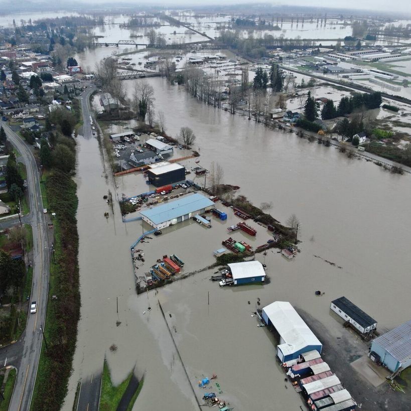 Aerial view of flooding affecting an industrial area with partially submerged buildings, vehicles, and equipment amidst a nearby road and residential neighborhood.