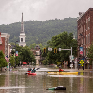 Street scene during a flood with two people kayaking, vehicles stranded, and flooded buildings in a small town with a church steeple and forested hills in the background.