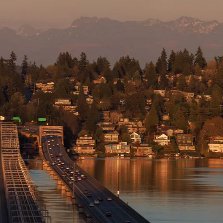 A bridge over a body of water with houses on a hillside and mountains in the background at sunset.