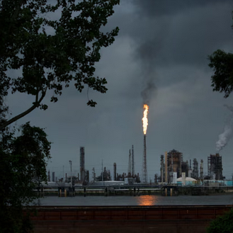 Industrial factory with smokestacks emitting smoke and a large flare burning in the sky during cloudy weather.
