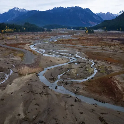 A winding river flows through a dry, barren landscape with distant mountains and a cloudy sky in the background.