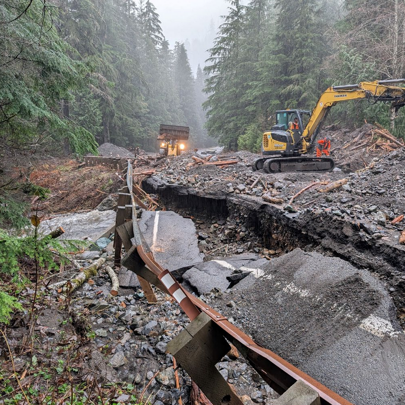 Construction crews repairing a damaged railway track after a landslide in a forest, with an excavator working on the debris and a dump truck in the background.