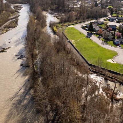 Aerial view of a river with partially frozen water and leafless trees along the bank. Nearby are residential houses with a grassy area and a pathway.