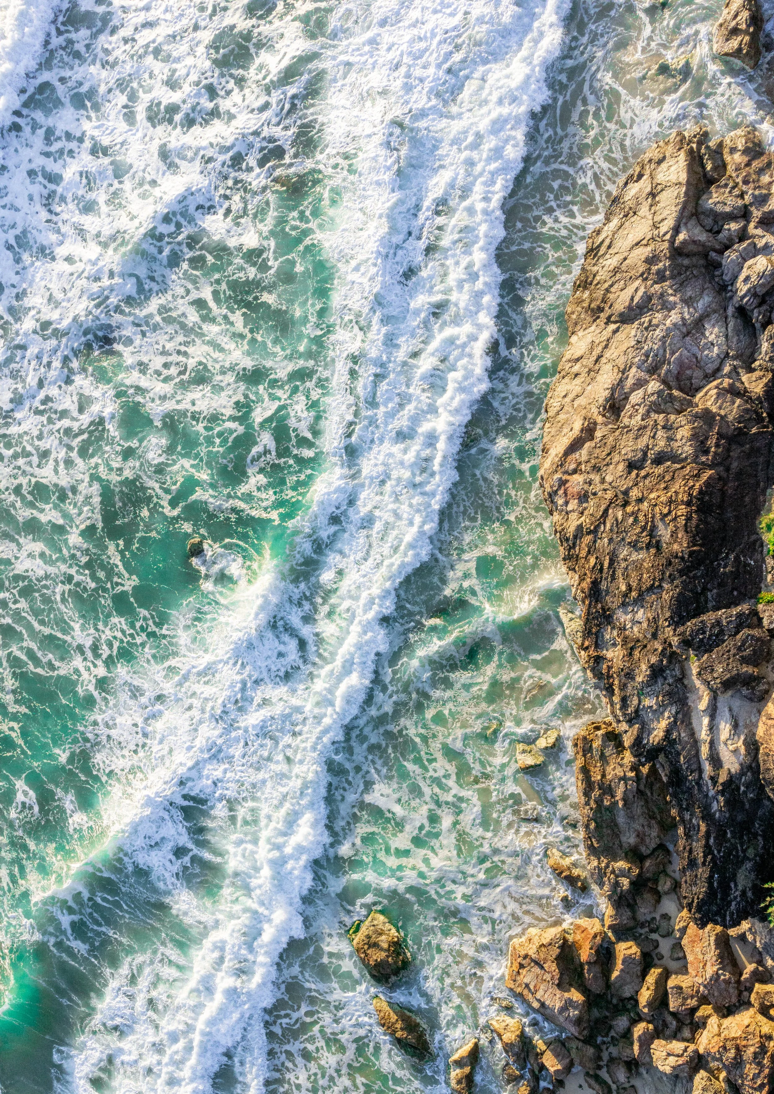 Aerial view of ocean waves crashing against rocky shoreline with white foam and turquoise water.