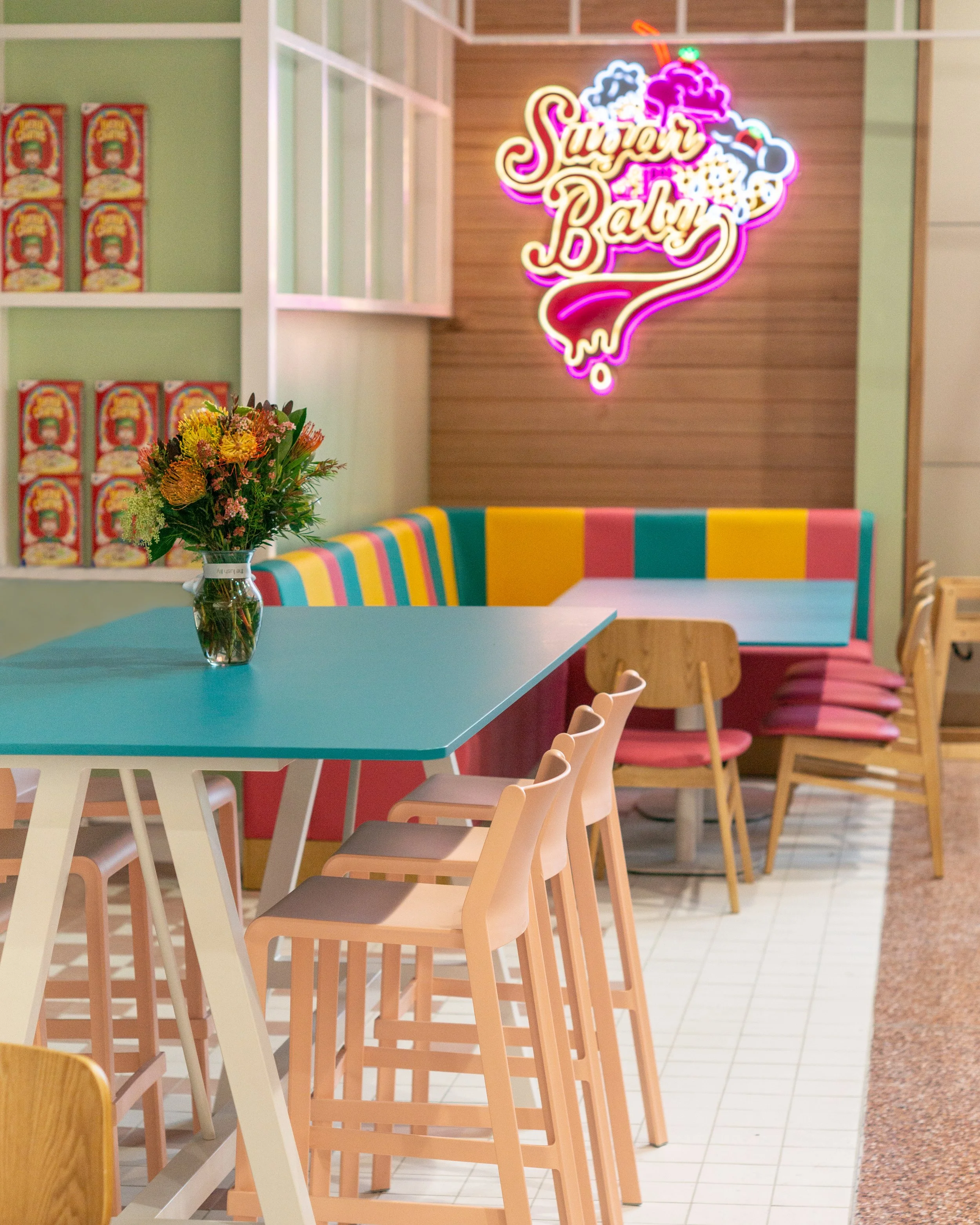Colorful ice cream-themed neon sign that says 'Sugar Baby' on a wooden wall behind a pink, teal, yellow, and multicolored booth seating. A vase with flowers on a turquoise table and pastel-colored chairs in a cafe interior.