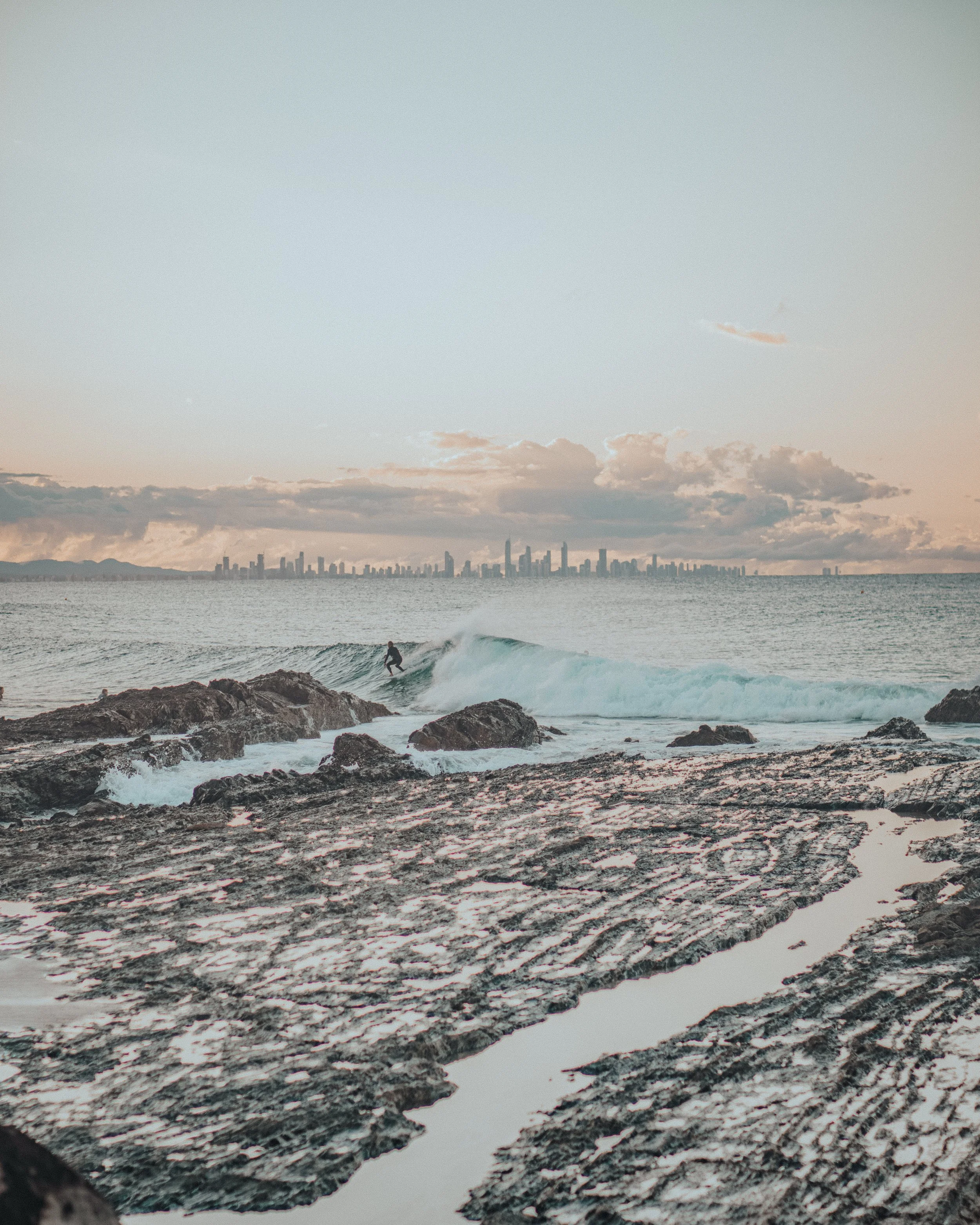 A surfer riding a wave near rocky shoreline with city skyline in the background at sunset.