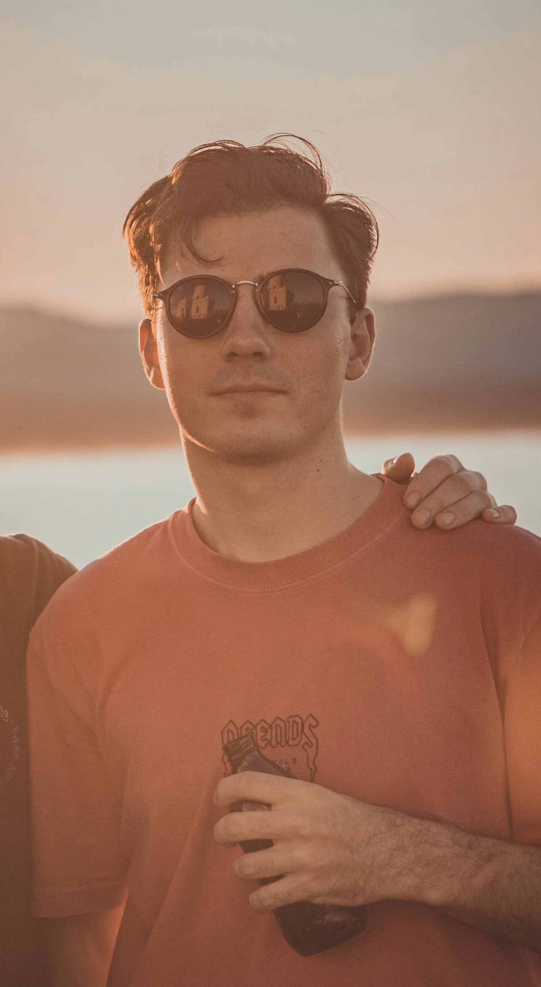 A young man with short brown hair wearing round sunglasses and an orange T-shirt, holding a dark bottle, outdoors during sunset, with someone else’s hand on his shoulder.