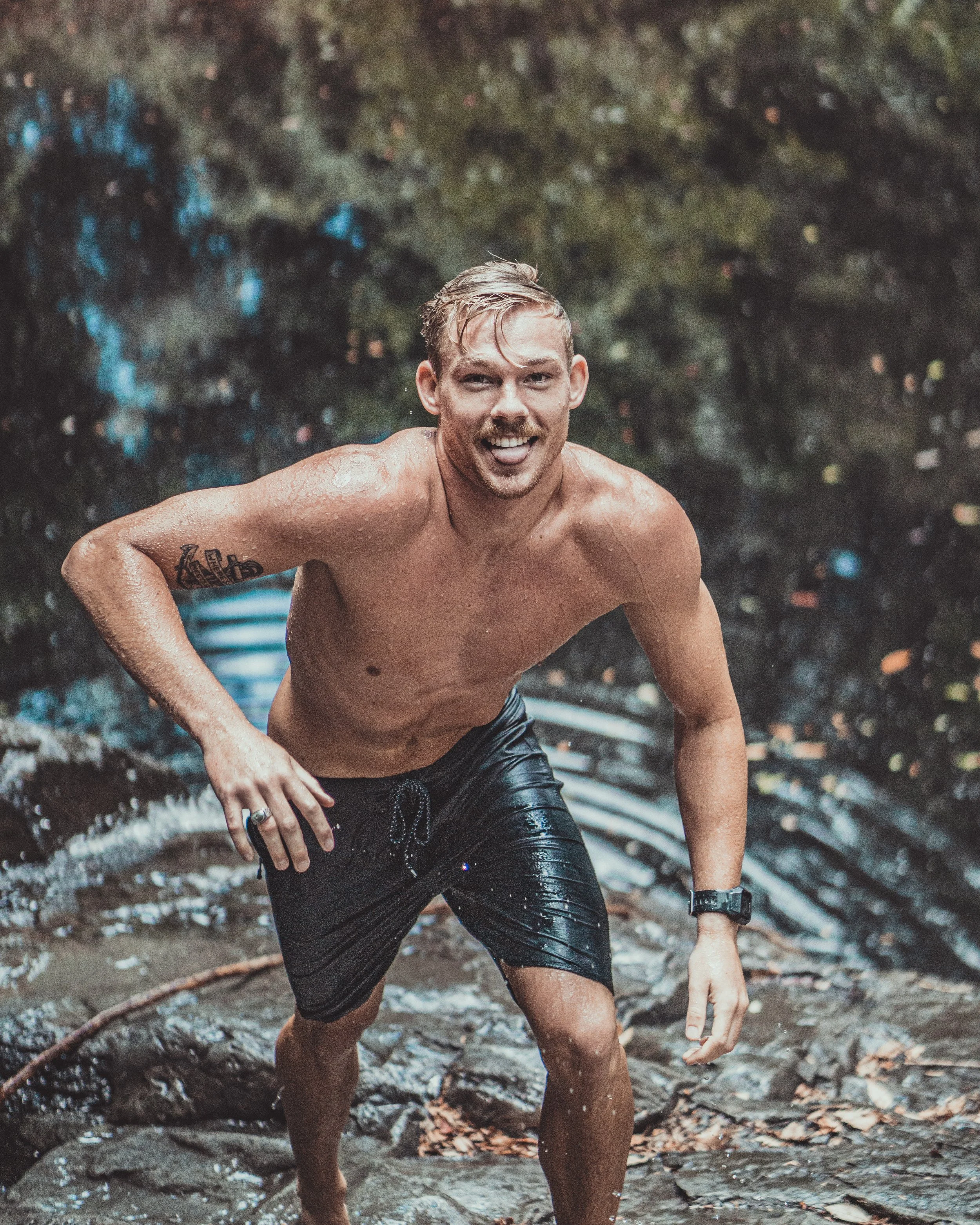 A man smiling and walking up waterfall surroundings, shirtless and wearing black shorts, with water on his body and a tattoo on his left arm.