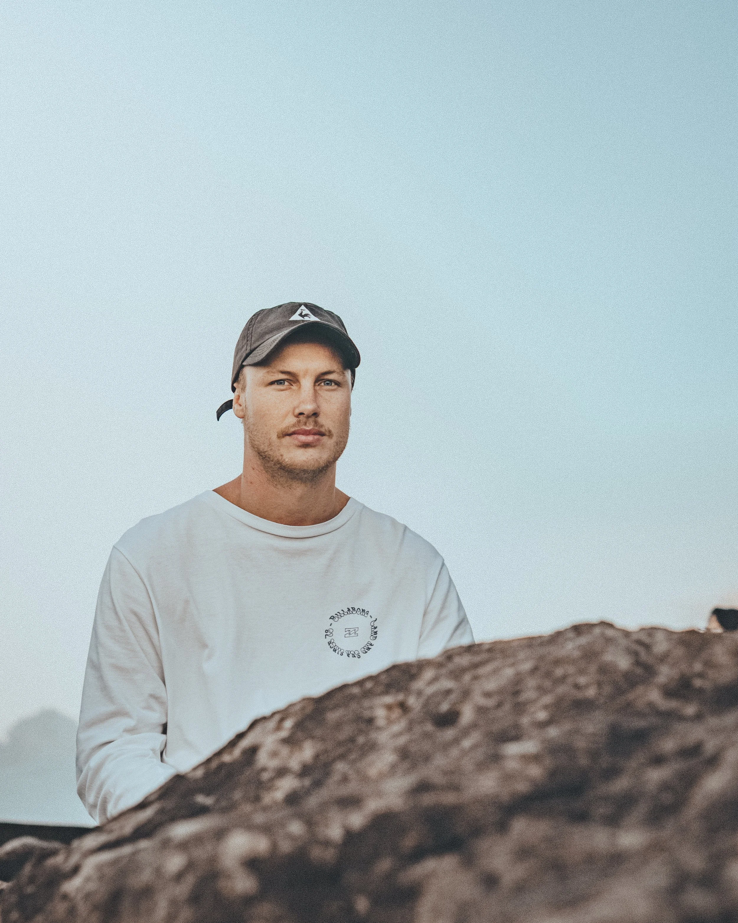 A man in a white long sleeve shirt and black cap standing outdoors near rocks with a clear sky in background. At Coolangatta
