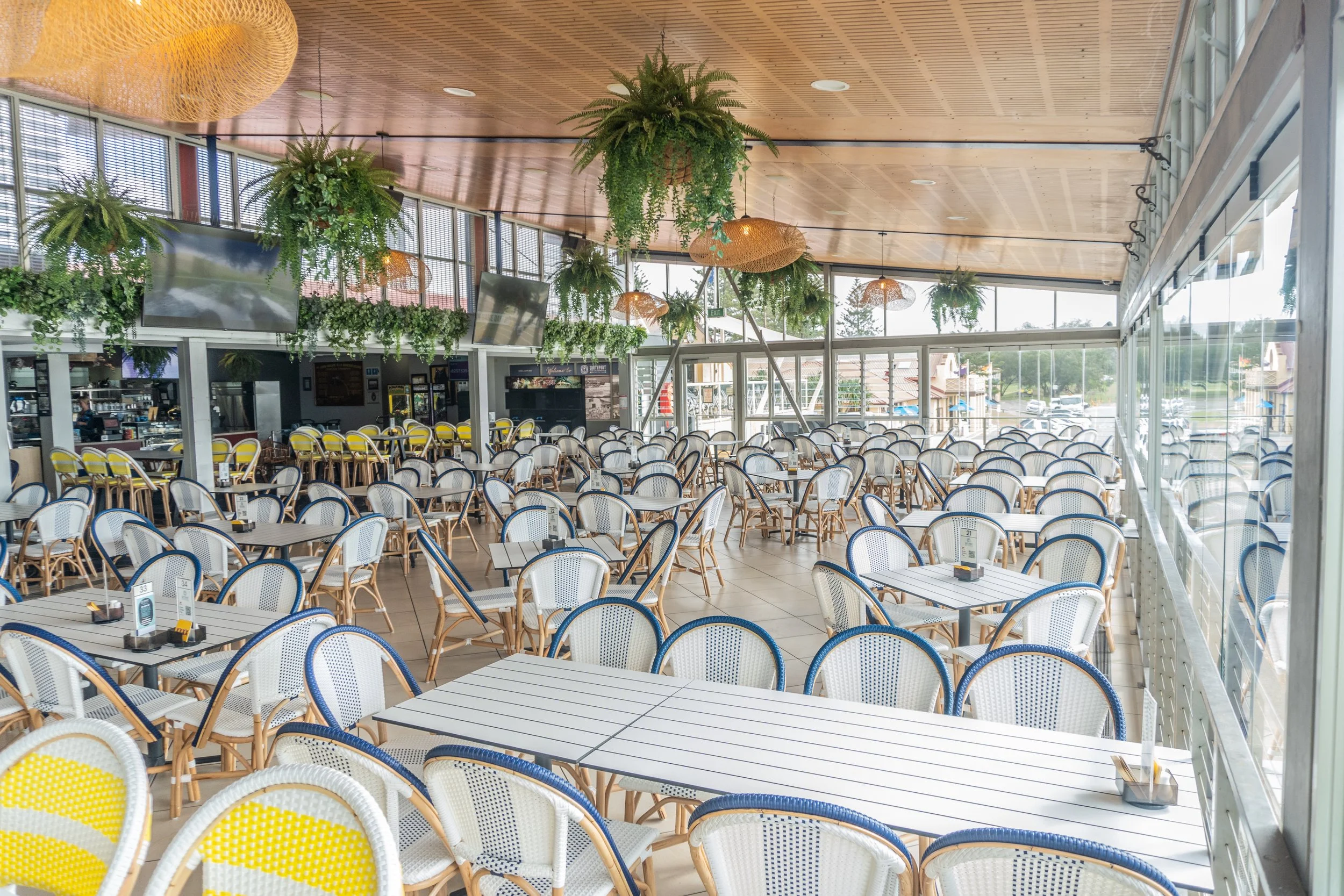 Empty restaurant with white tables and chairs, hanging plants, and large windows letting in natural light.