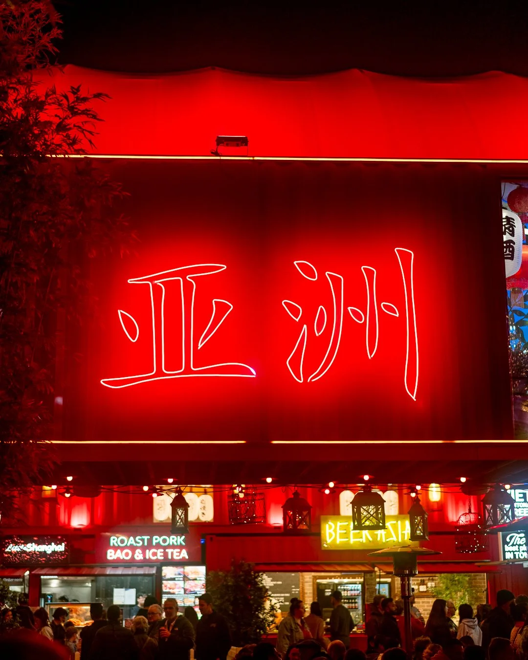 Large red neon sign with Chinese characters above a busy food stall at night, serving roast pork, bao, and ice tea, with a crowd of people and hanging lanterns.