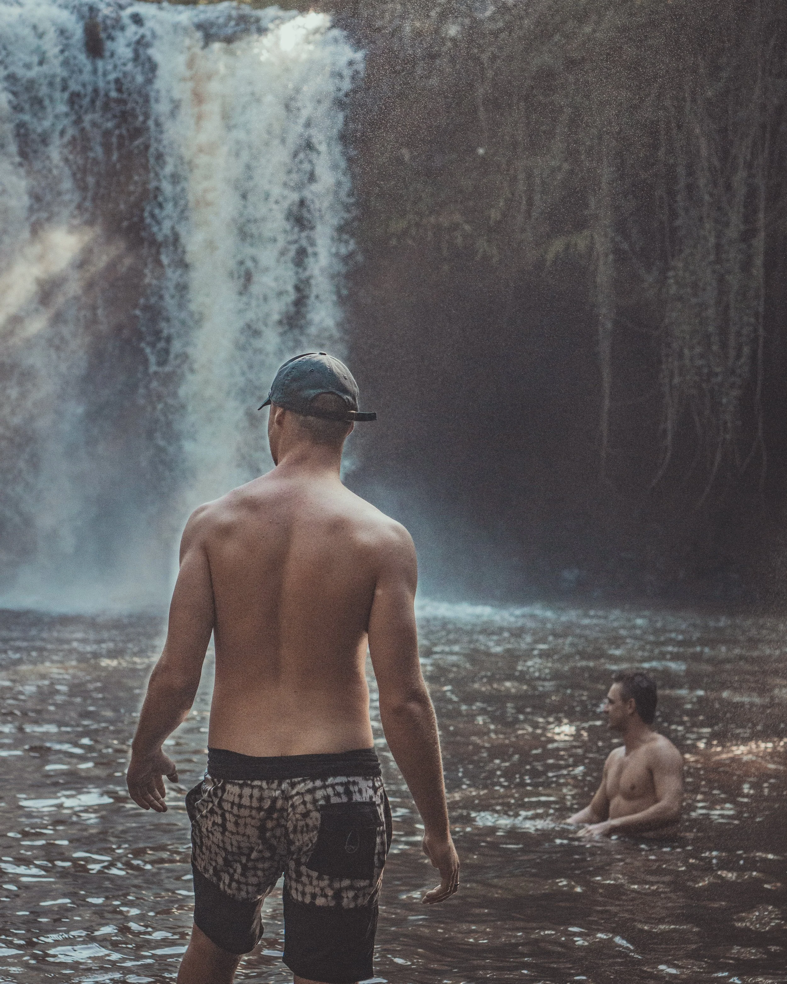 Two shirtless men in swim trunks enjoy a waterfall in a natural setting, one standing in the water and the other sitting deeper in the pool, with a large waterfall cascading in the background.