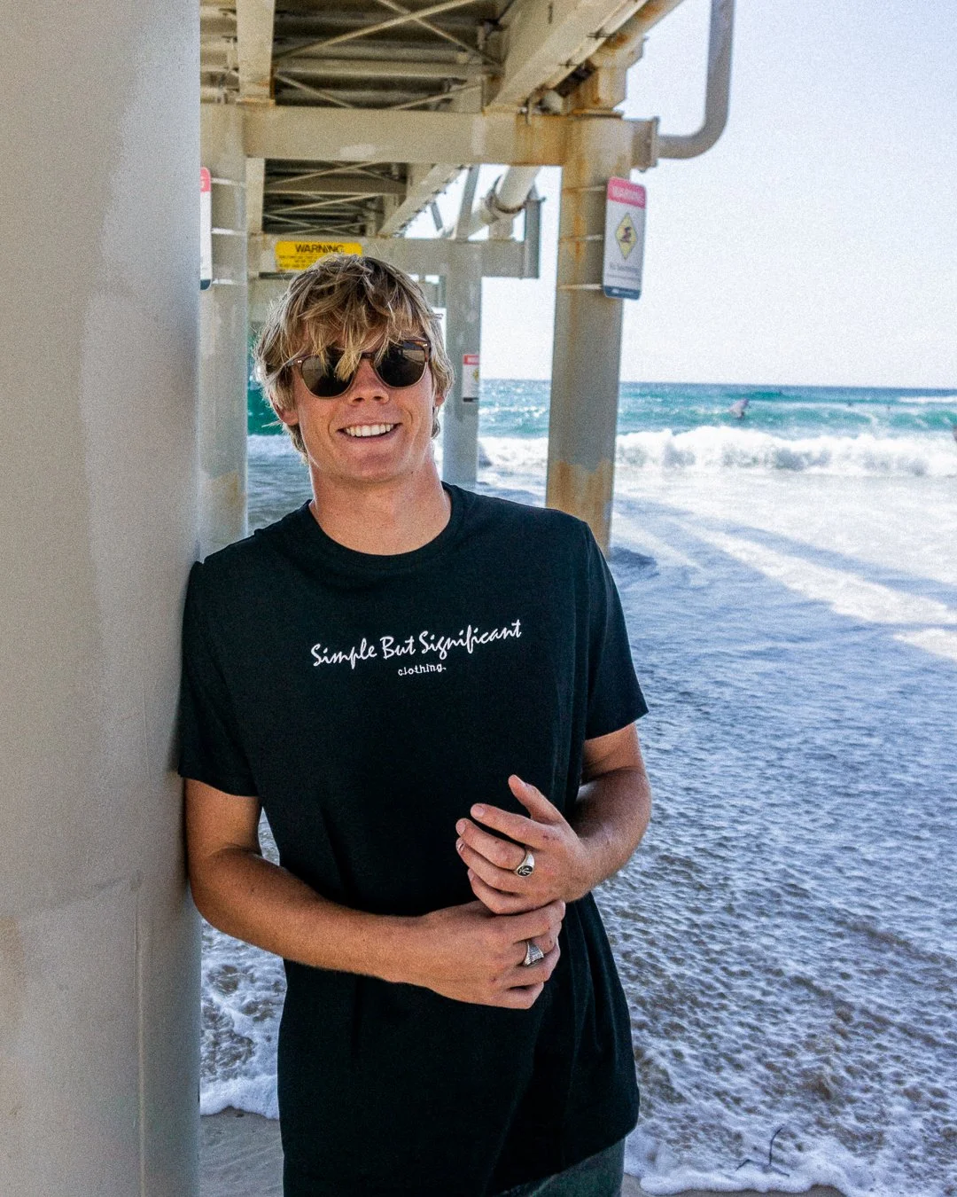Young man with wavy blonde hair wearing sunglasses and a black T-shirt with white text, standing under a pier on a beach with ocean waves in the background.