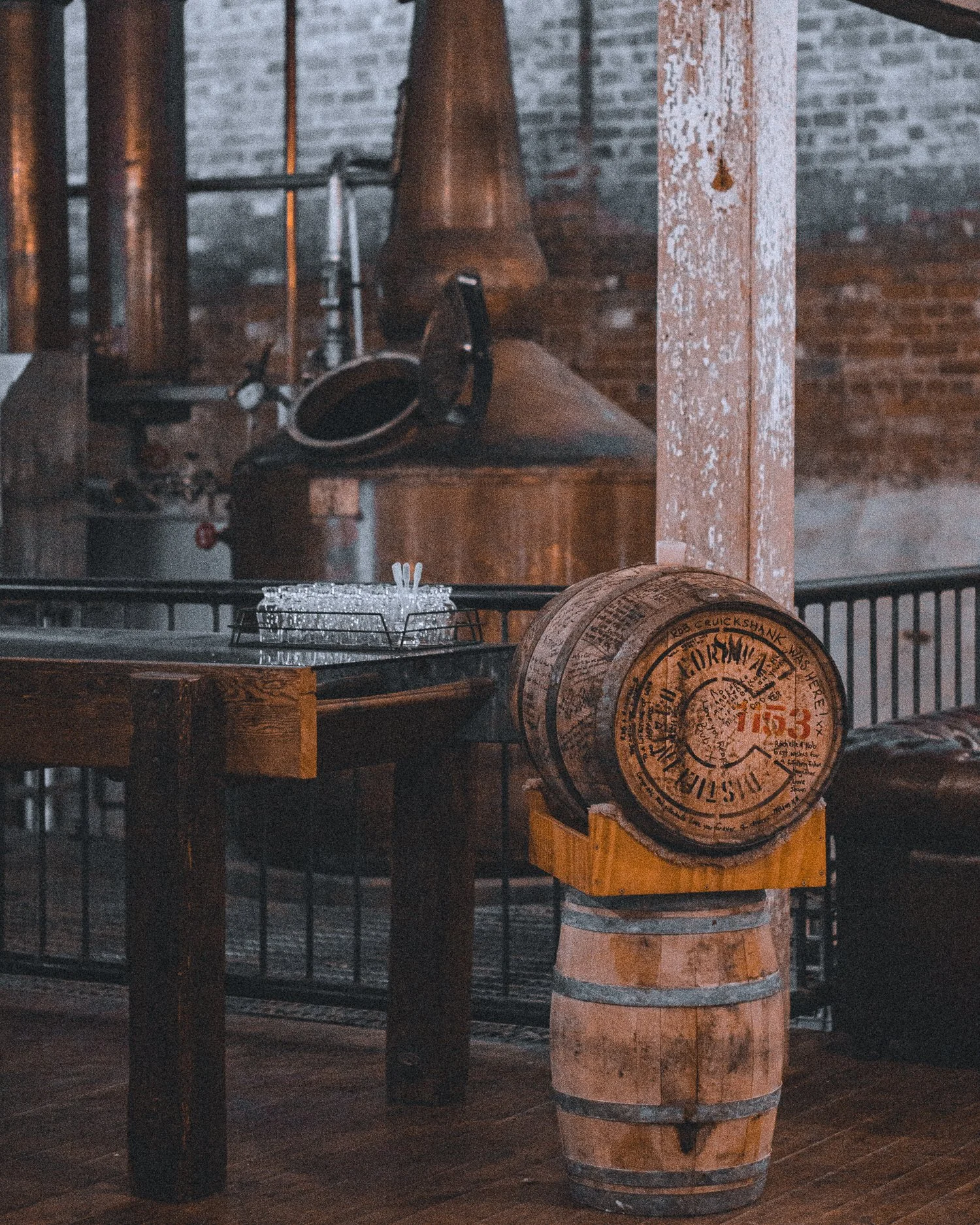 A rustic interior with a wood barrel resting on a wooden stand, a round metal container on top, and a large copper brewing kettle in the background, all set against brick walls. At Corowa Whiskey and Chocolate (Corowa Distilling Co)