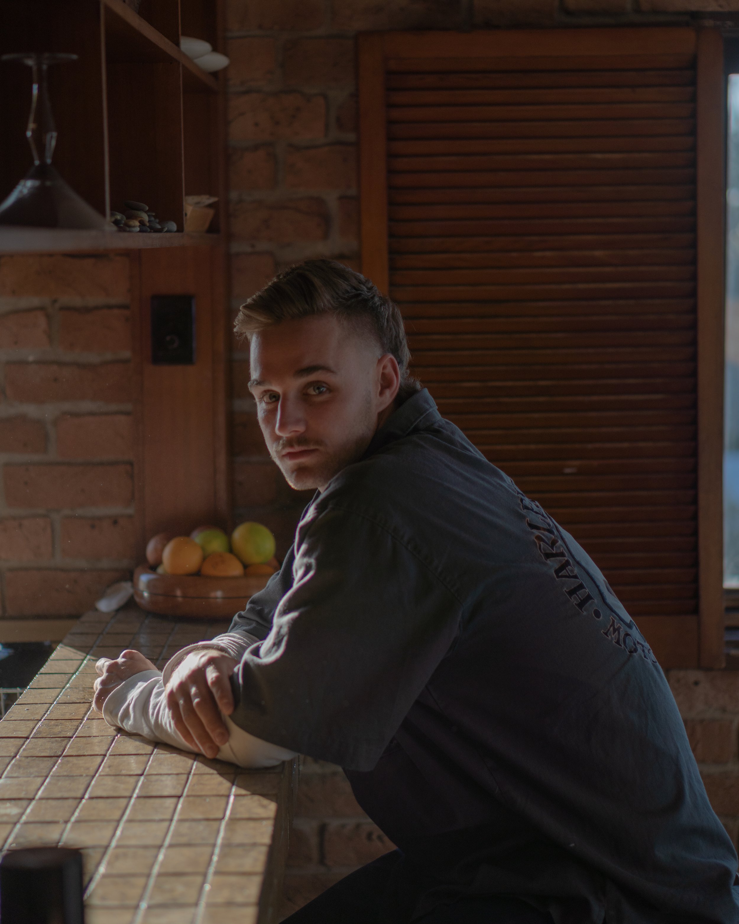 A young man with styled hair sitting at a tiled kitchen counter, with a brick wall behind him and a bowl of mixed fruits beside him.