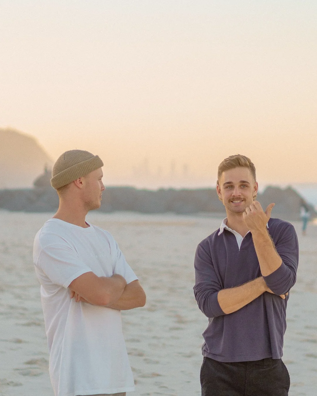 Two young men standing on a beach at sunset, one is wearing a white t-shirt and beanie, with arms crossed, looking at the other, who is in a navy polo shirt, giving a thumbs-up and smiling. At Gold Coast