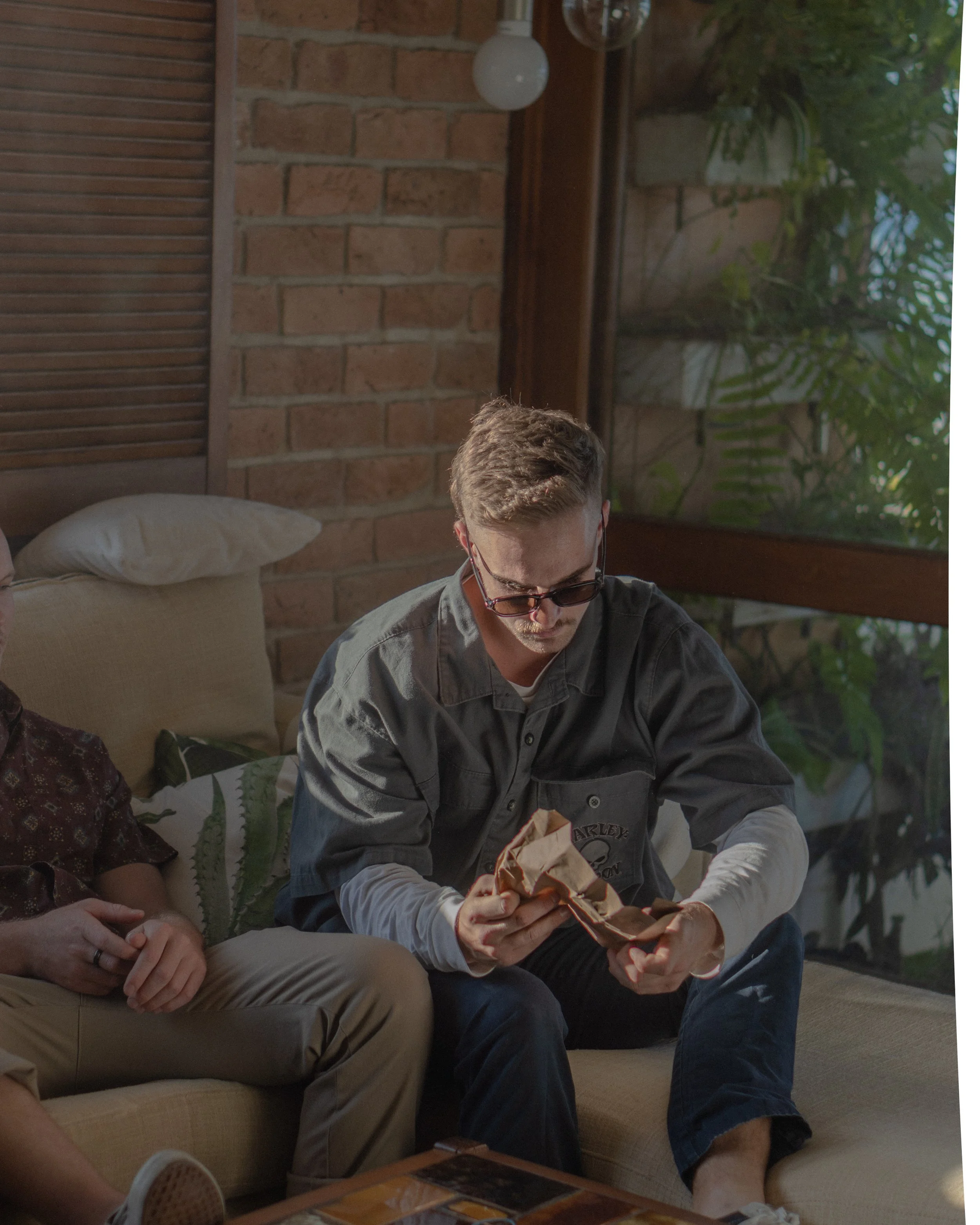 A young man with glasses sits on a beige sofa, looking at a crumpled paper bag in his hands in a cozy room with a brick wall and window behind him.