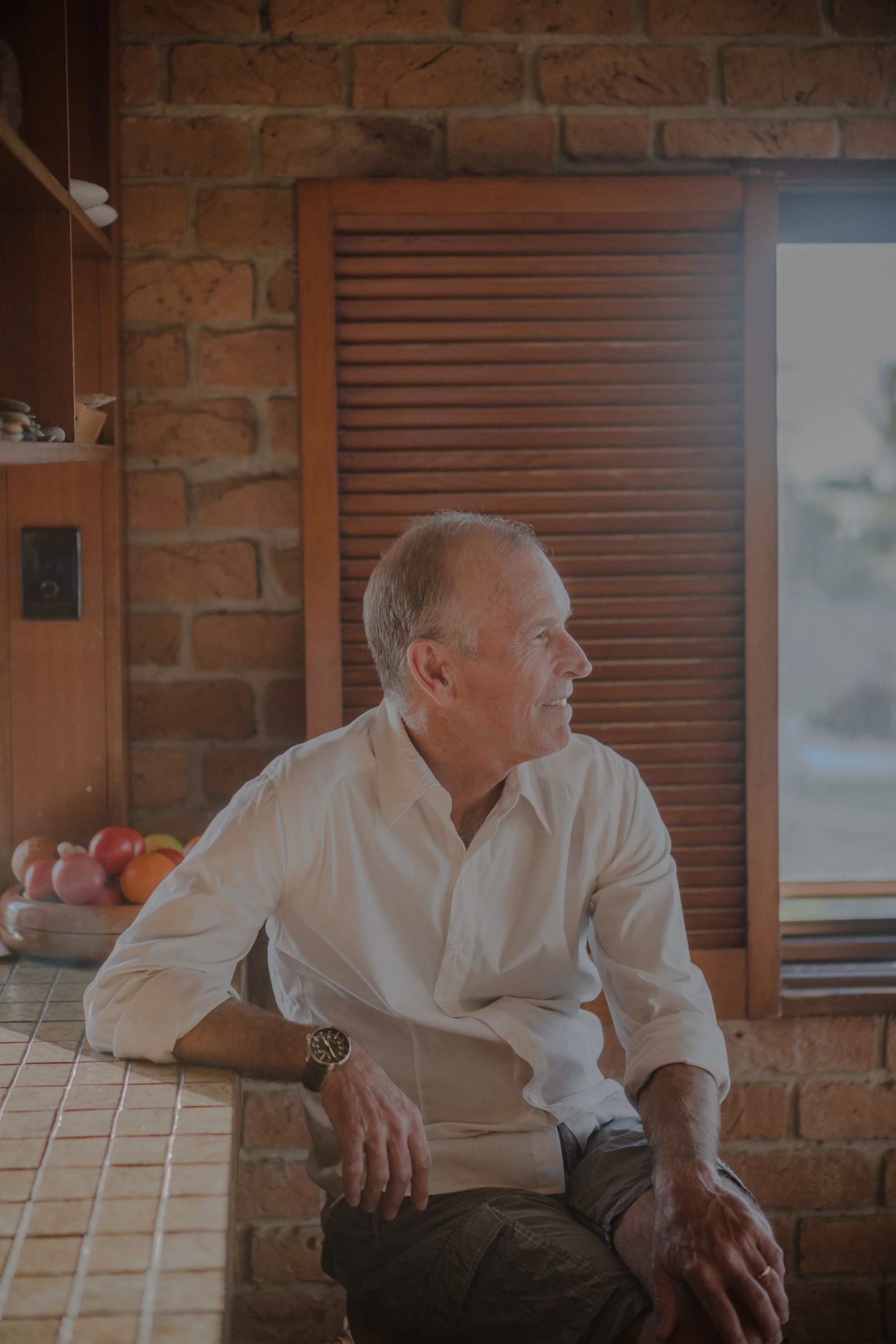 A middle-aged man sitting at a kitchen counter, looking out the window, smiling. He has short, light-colored hair, is wearing a cream-colored shirt and a watch.