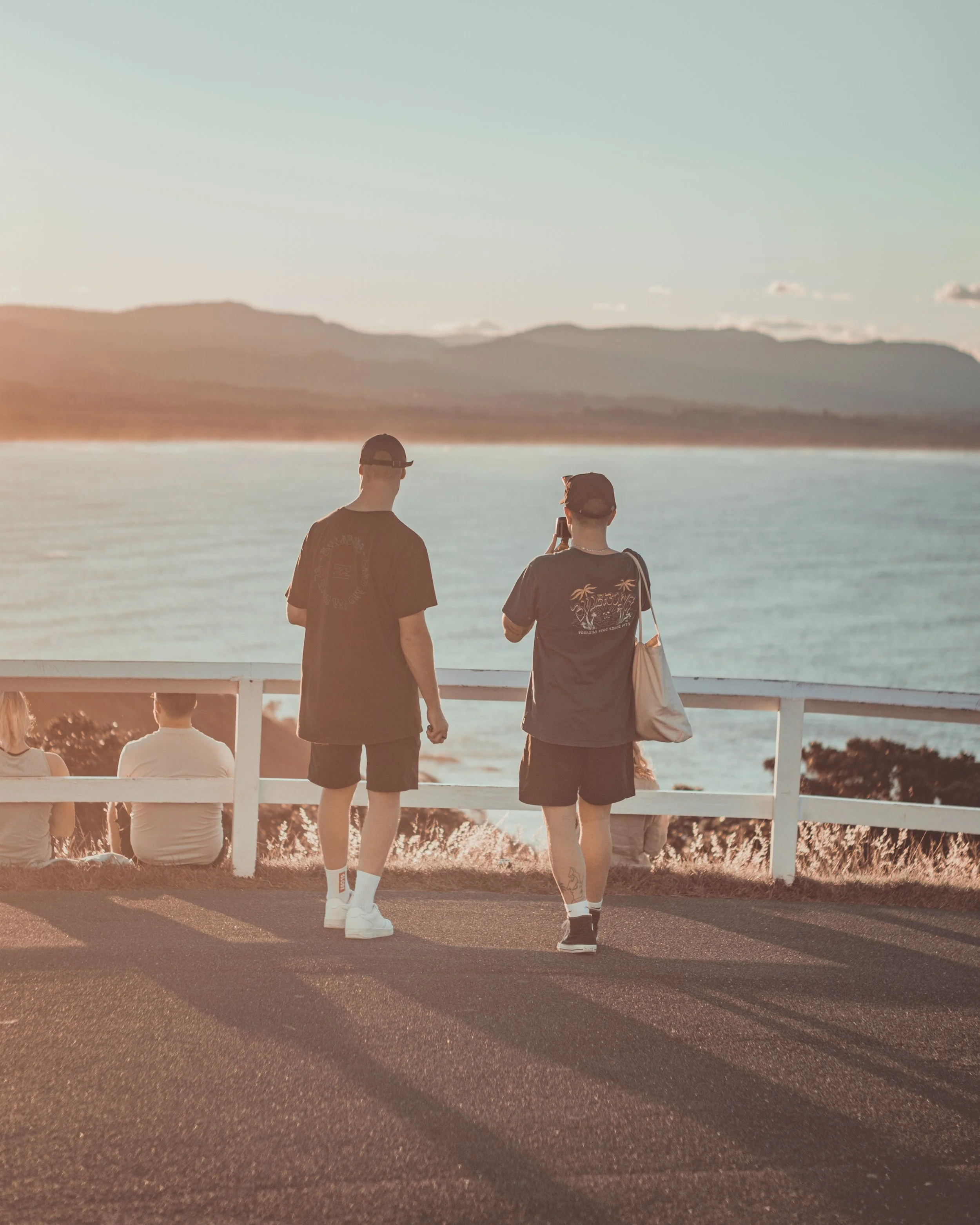 Two young men walking near a railing by a large body of water during sunset, with other people sitting and relaxing nearby. At Byron Bay