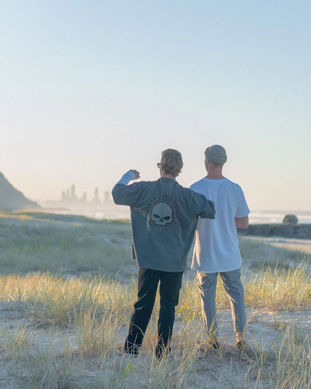 Two men stand on the beach facing the ocean at sunset. One wears a Harley Davidson jacket with a skull logo, the other a white t-shirt and gray beanie.