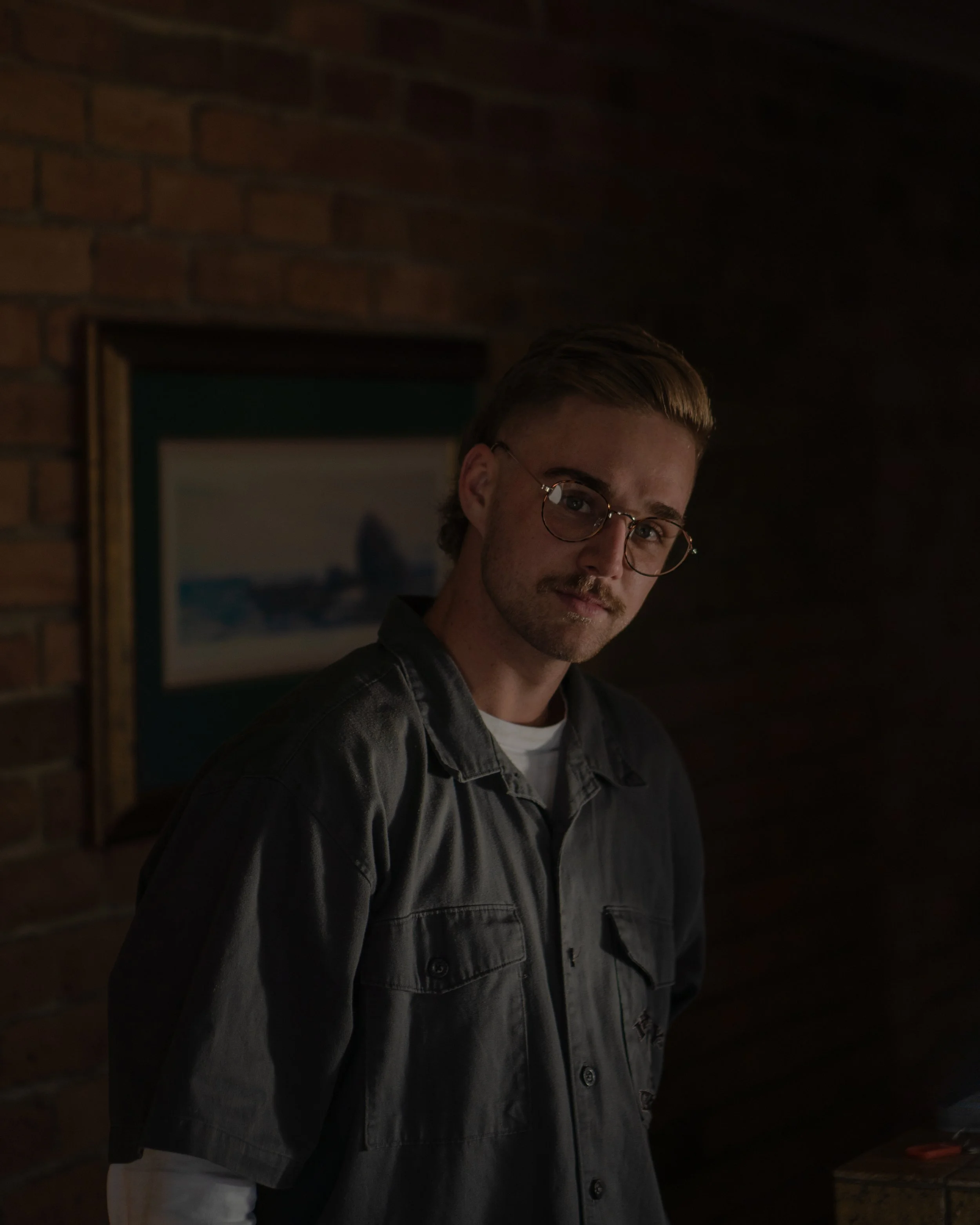A young man with light-colored hair, glasses, and a mustache, wearing a dark gray shirt over a white long-sleeve shirt, standing in a dimly lit room with a brick wall and a framed painting in the background.