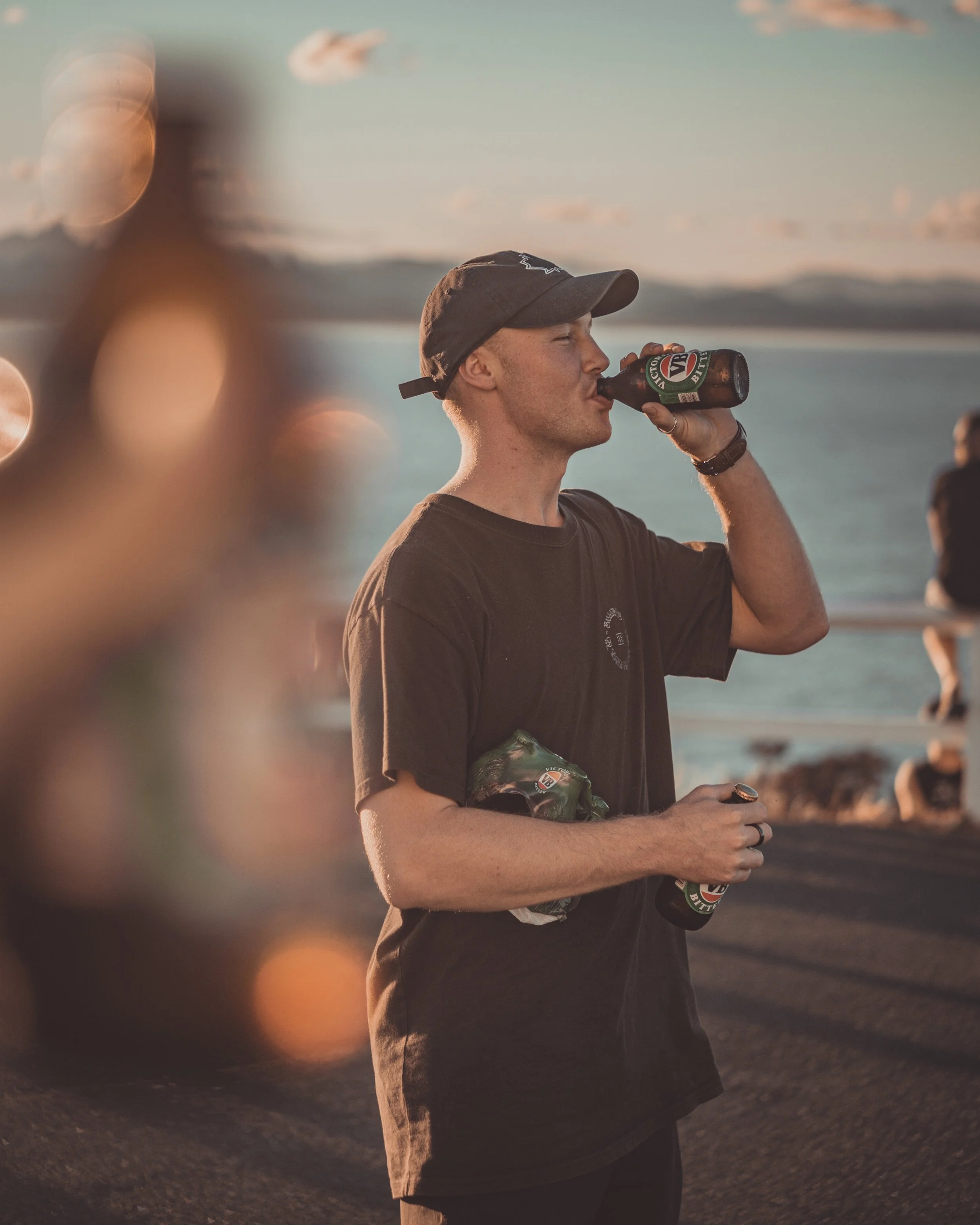 A young man in a dark t-shirt and baseball cap drinking a beer from a brown bottle, with other people in the background near a body of water at sunset. At Byron Bay