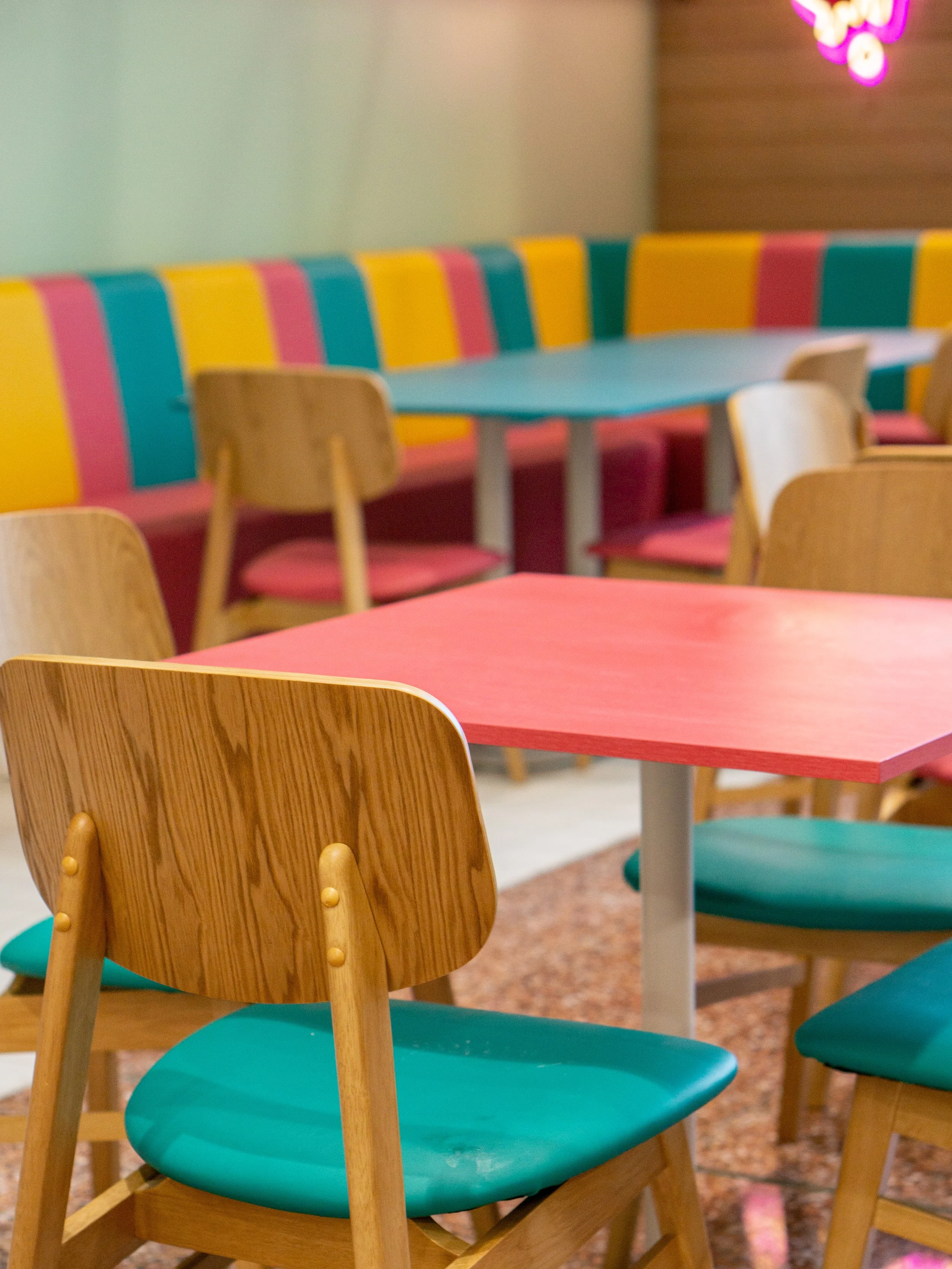 Empty colorful restaurant dining area with wooden chairs, a red table, a pastel teal table and a multicolored, striped booth in the background.