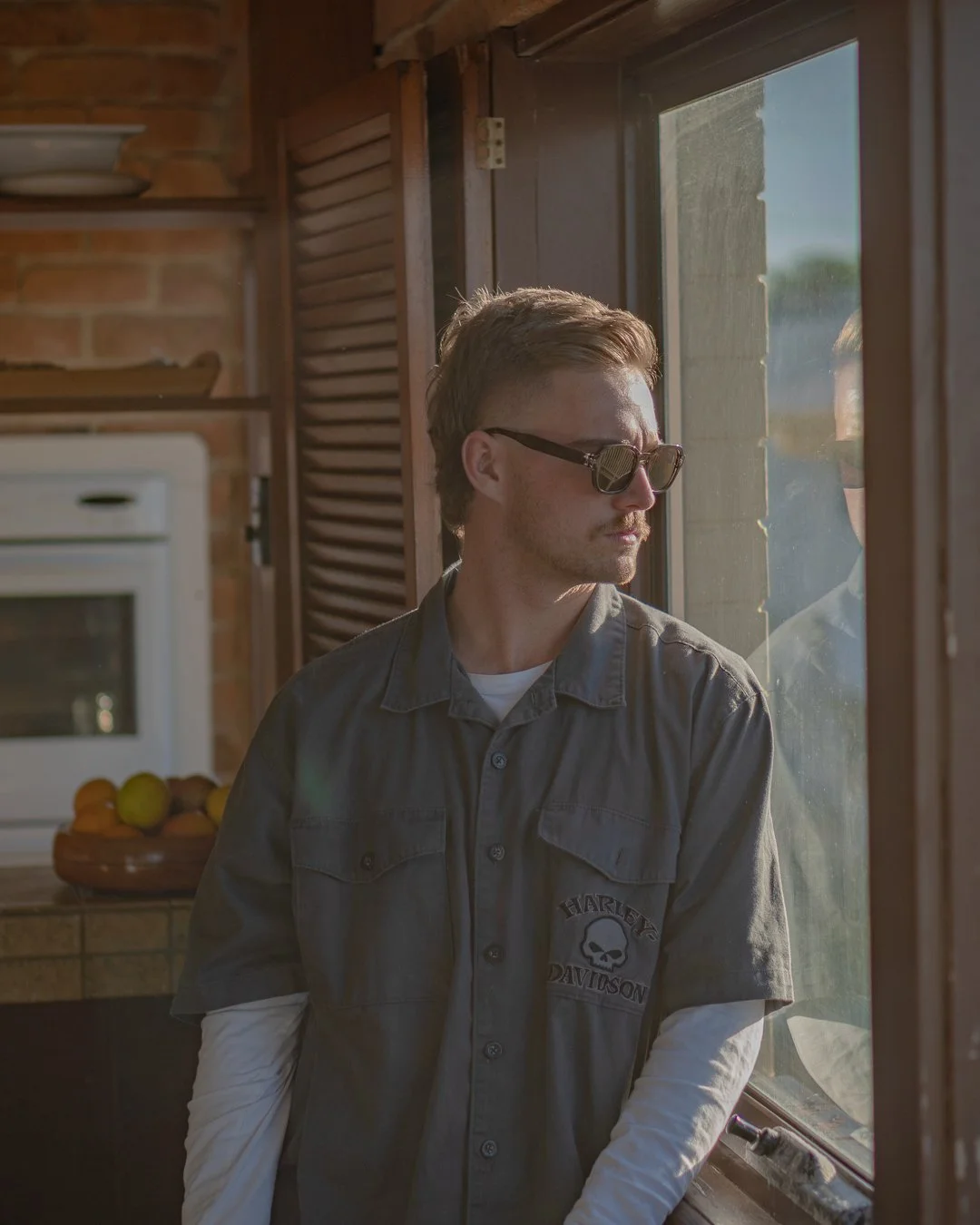 A young man with light brown hair, wearing sunglasses and a Harley Davidson shirt, stands inside near a window, looking outside with sunlight illuminating his face.
